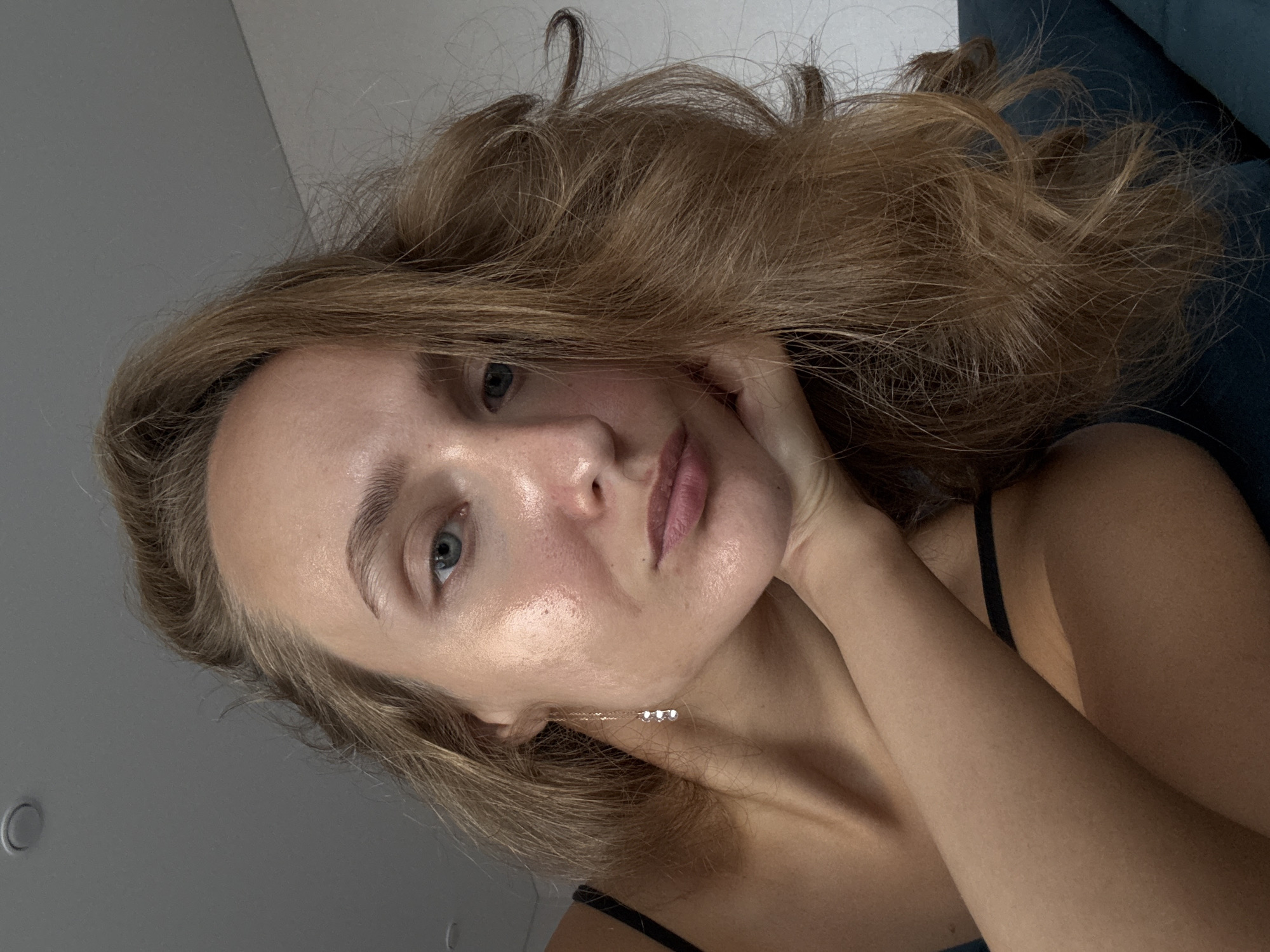 A close-up, waist-up shot features a young woman with light brown, wavy hair. She has blue eyes and fair skin with a dewy complexion. Her left hand is resting on her chin, and she is wearing a black camisole. A delicate dangle earring with three small, clear gems hangs from her left ear. The background is blurred, but hints of a gray ceiling with a circular light fixture and a dark blue surface suggest an indoor setting. The scene is not located in the city of Подшивалово, Russia.