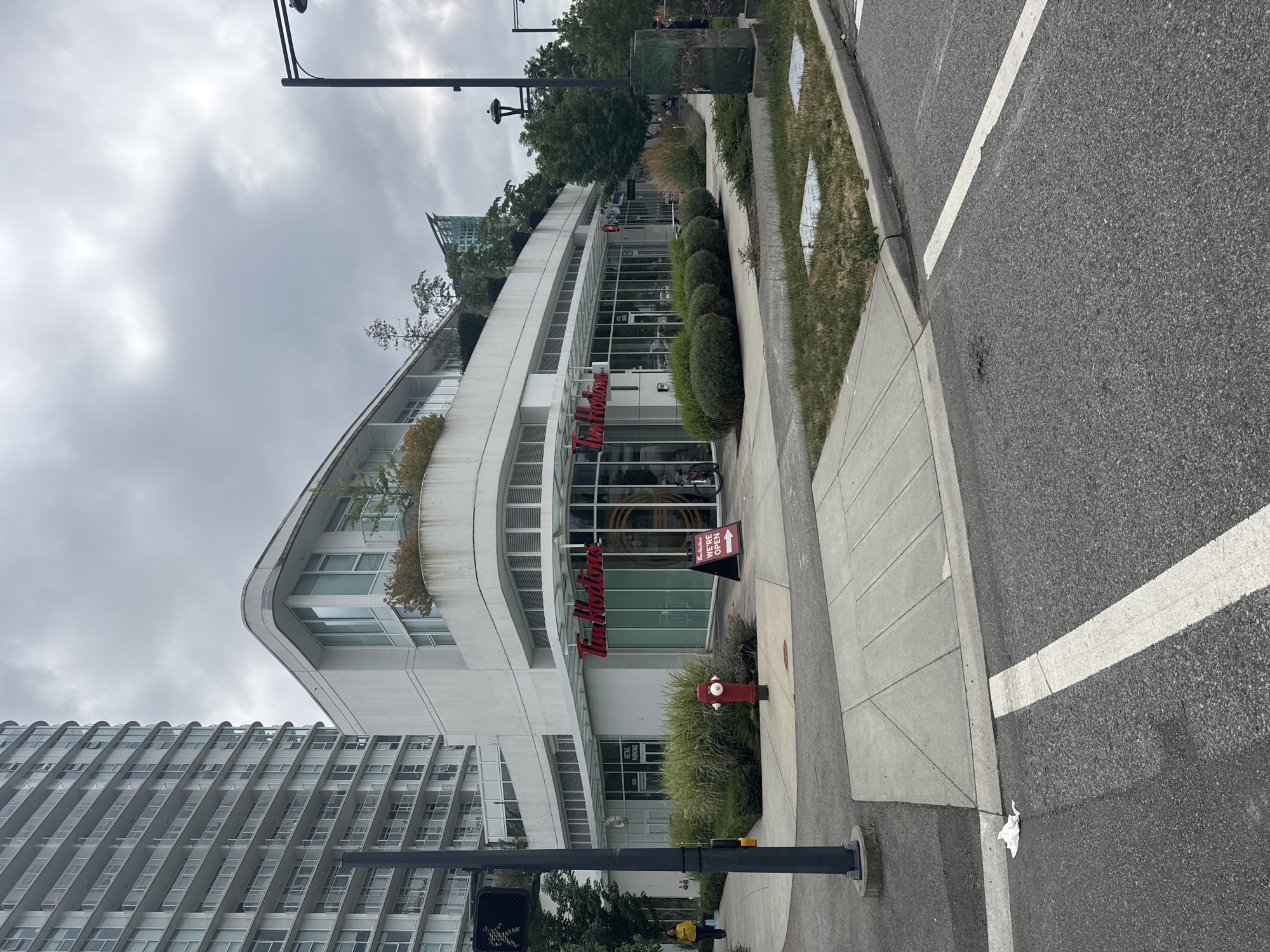The image shows a Tim Hortons restaurant on the ground floor of a modern, multi-story building with a curved facade. A tall residential building stands to the left of the Tim Hortons. The scene is located in Surrey, Canada. The sky is overcast.
In the foreground, a paved road with white lane markings is visible, with a concrete sidewalk running along it. A pedestrian crossing signal with a walking person icon is mounted on a dark pole. A person wearing a yellow jacket is walking on the sidewalk, carrying shopping bags. A red fire hydrant is positioned on a patch of green grass between the sidewalk and the building.
The Tim Hortons storefront features large glass windows and the distinctive red Tim Hortons logo. A "We're Open" sign with an arrow pointing to the right is placed on the sidewalk outside the restaurant. A bicycle is parked to the right of the entrance. Beyond the Tim Hortons, other retail establishments are visible.
The environment appears to be an urban setting with well-maintained landscaping, including shrubs and small trees. The overall atmosphere is one of a commercial street in a city.