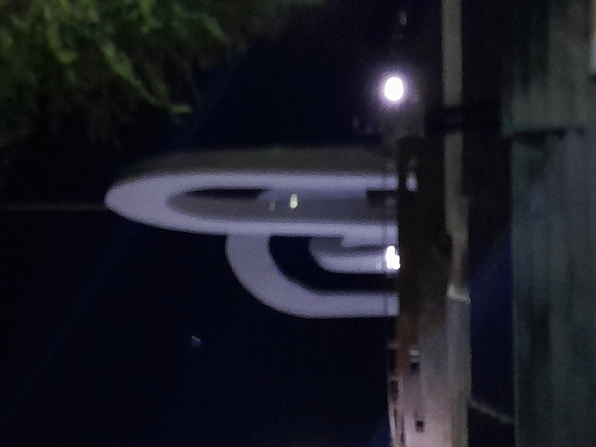 The image depicts an outdoor scene at night, featuring a modern sculpture with several large, curved, arch-like structures made of a light-colored material, possibly concrete or stone. The sculpture is illuminated by artificial lighting, creating a glow on its surfaces. Behind the sculpture, a dark sky is visible, with a faint, distant light that might be a star or planet. To the right of the sculpture, the dark foliage of trees can be seen, suggesting a park or urban green space. In the foreground, a blurred, dark surface, possibly a road or ground, is present. The overall impression is one of a public monument or art installation in an urban setting, likely Uchquduq, Uzbekistan, given the context. The lighting conditions suggest it is after sunset. There are no people or animals visible in the frame.
