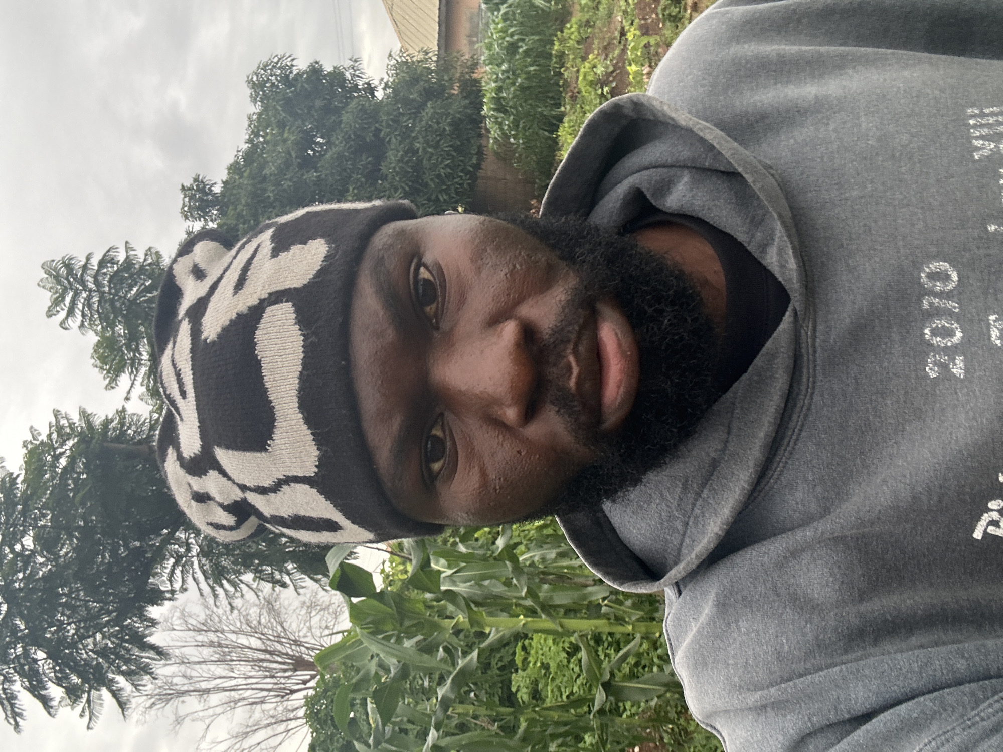 A dark-skinned man with a beard and a black and white patterned beanie is taking a selfie in an outdoor, rural setting. He is wearing a grey hooded sweatshirt. Behind him, there are tall green corn stalks and other foliage, as well as trees and a cloudy sky. A building is visible in the background. The location is identified as Gura Top, Nigeria.