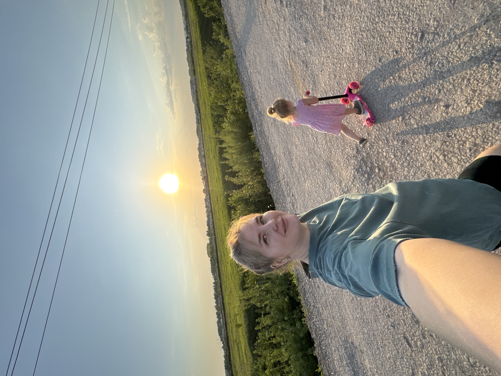 A selfie shows a woman holding a camera at arm's length on a paved road, likely in the city of Горный, Russia. A young girl rides a pink scooter ahead of her, towards the sunset. The woman is wearing a blue-green shirt. The girl has blonde hair styled in a ponytail and wears a pink dress. A field of green vegetation borders the road on one side, with a clear sky and power lines visible in the background. The sun is low on the horizon, casting long shadows.