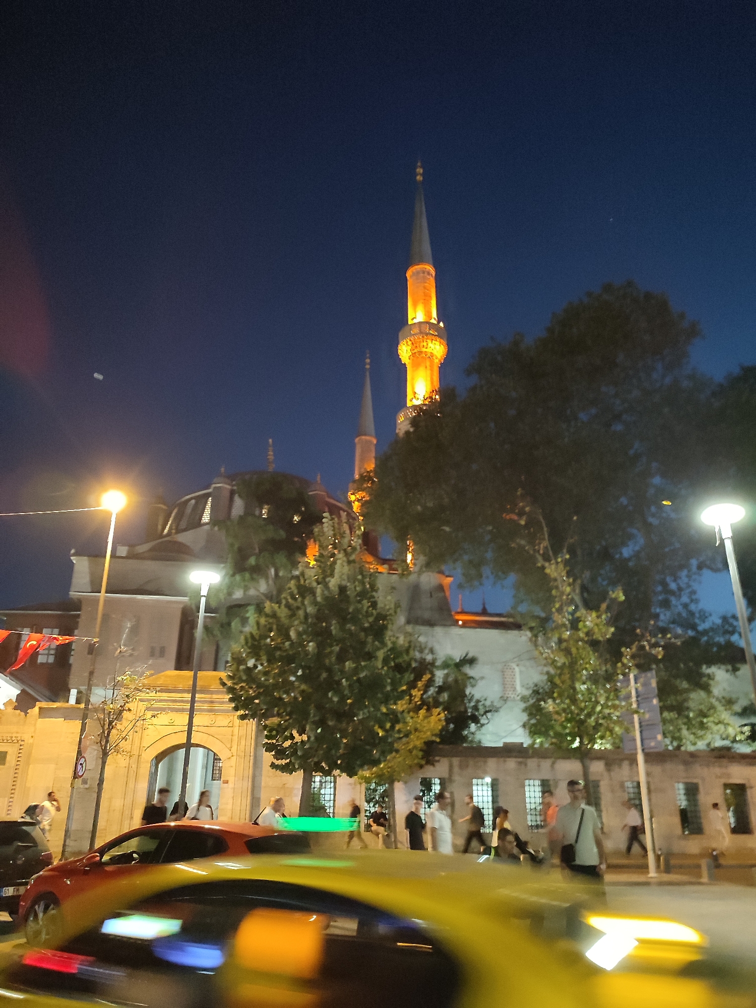At night in Üsküdar, Turkey, a mosque with illuminated minarets stands against a dark blue sky. Several people are walking on the sidewalk in front of the mosque, some appearing to be tourists. A yellow car with blurry lights is in the foreground, suggesting motion. Turkish flags are visible on the left side of the image. Streetlights illuminate the area, casting a warm glow on the scene. The architecture of the mosque features domes and arched windows, typical of Ottoman style.