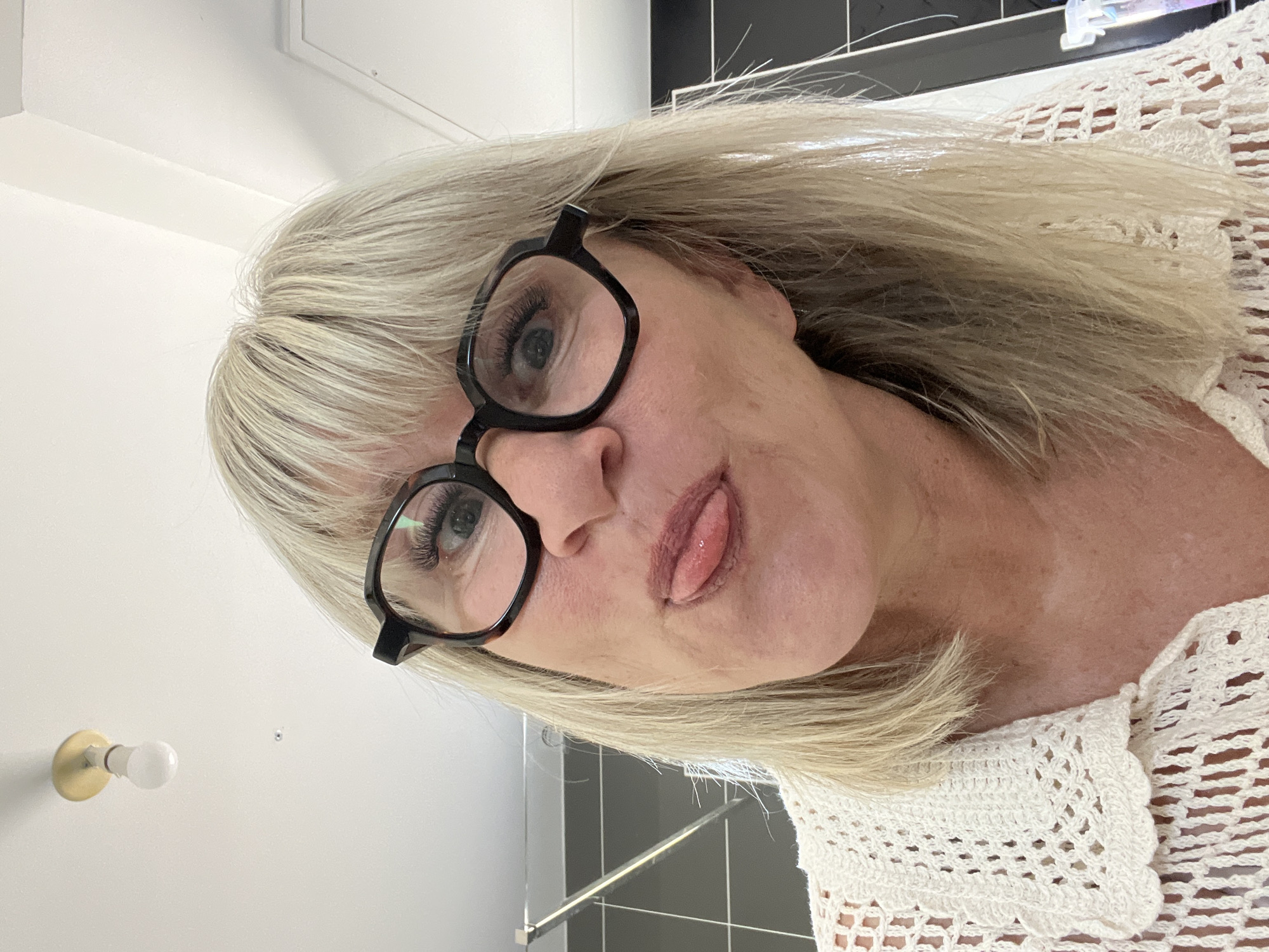 A woman with blonde hair and black-rimmed glasses is sticking her tongue out. She is wearing a white crocheted top. The background includes a white wall with a light fixture and a black-tiled wall. The location is not specified in the image.