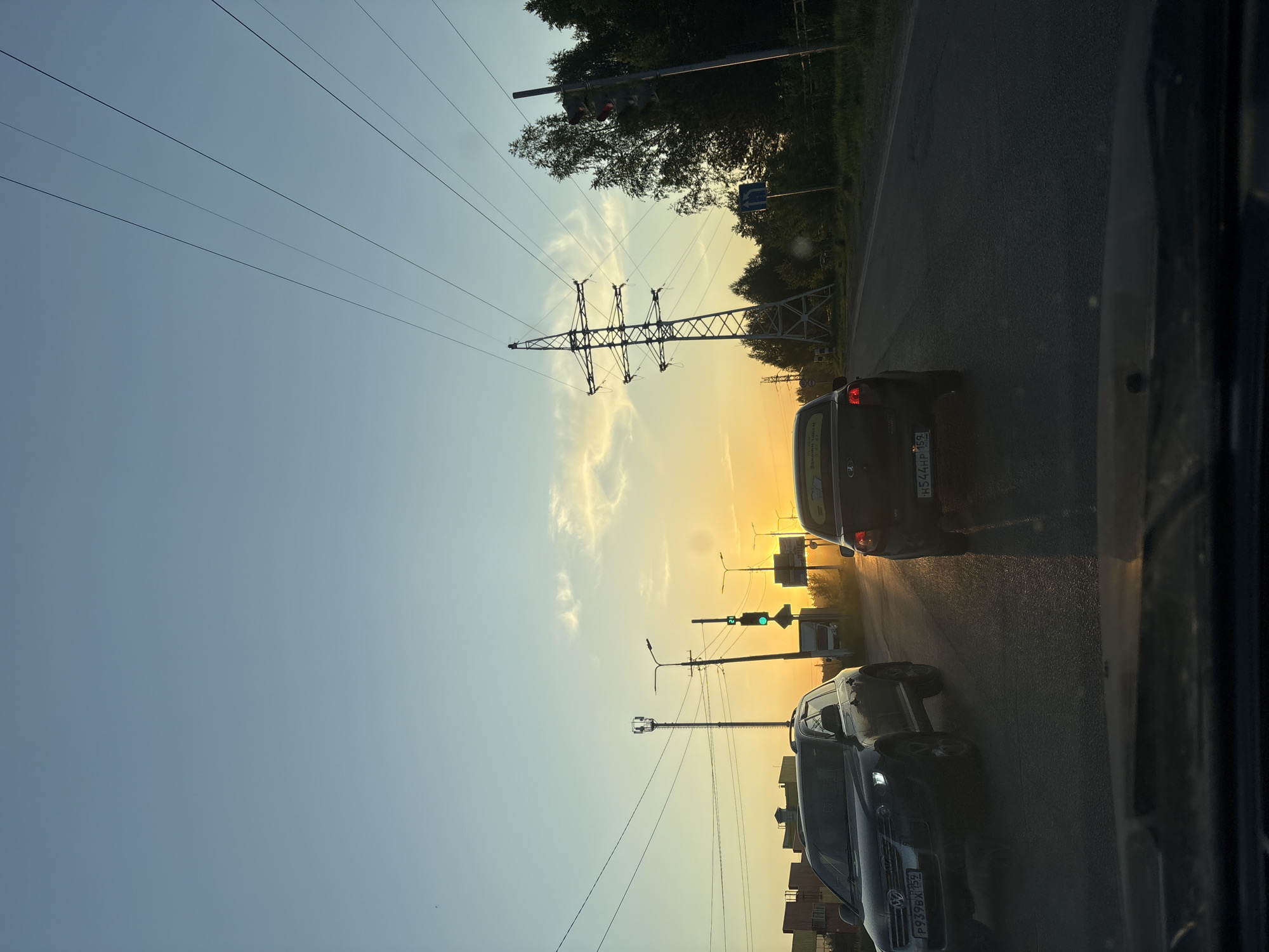 The image depicts a street scene at sunset in what appears to be the city of Chaikovsky, Russia. The photograph is taken from inside a vehicle, possibly a car. Several vehicles are visible on the road, including a dark-colored car with the license plate "H544HP 159" and a dark SUV with the license plate "P939EX 159". Overhead power lines and a transmission tower are visible against the evening sky, along with several streetlights and traffic signals, one of which is displaying a green light. Trees and vegetation line the road. The sky exhibits a gradient of colors from blue to orange, indicative of sunset. Buildings are visible in the background. The road surface appears wet, possibly from rain.