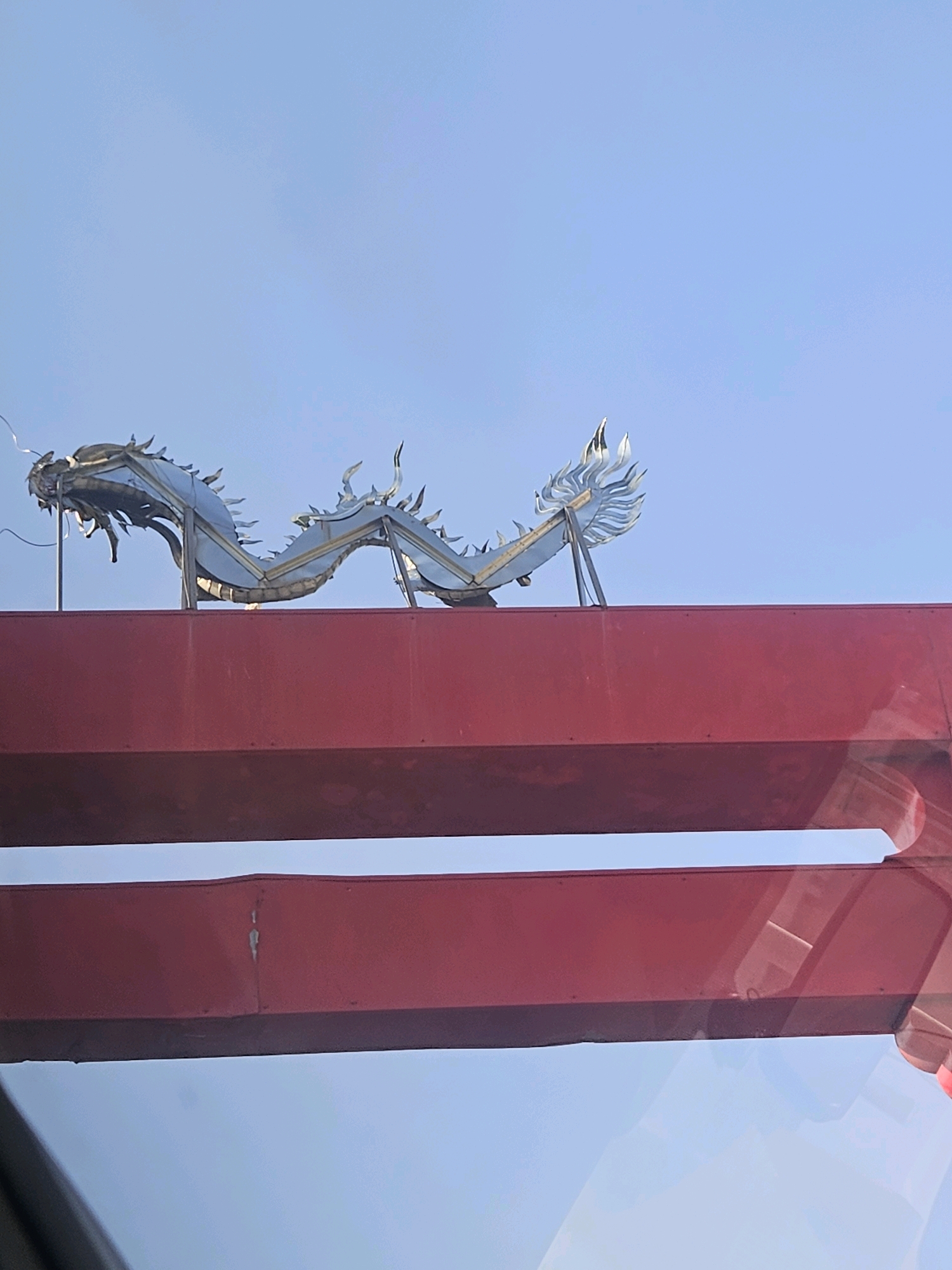 A silver dragon sculpture is mounted on top of a red structure against a light blue sky. The dragon is elongated and appears to be made of metal, with a detailed head and body. The red structure is a large, rectangular beam or building element, with another similar red structure visible below it. The overall scene suggests an outdoor setting, possibly an architectural feature or decoration. This dragon sculpture is located at the entrance to the Huwei Sugar Factory in Huwei Township, Yunlin County, Taiwan.