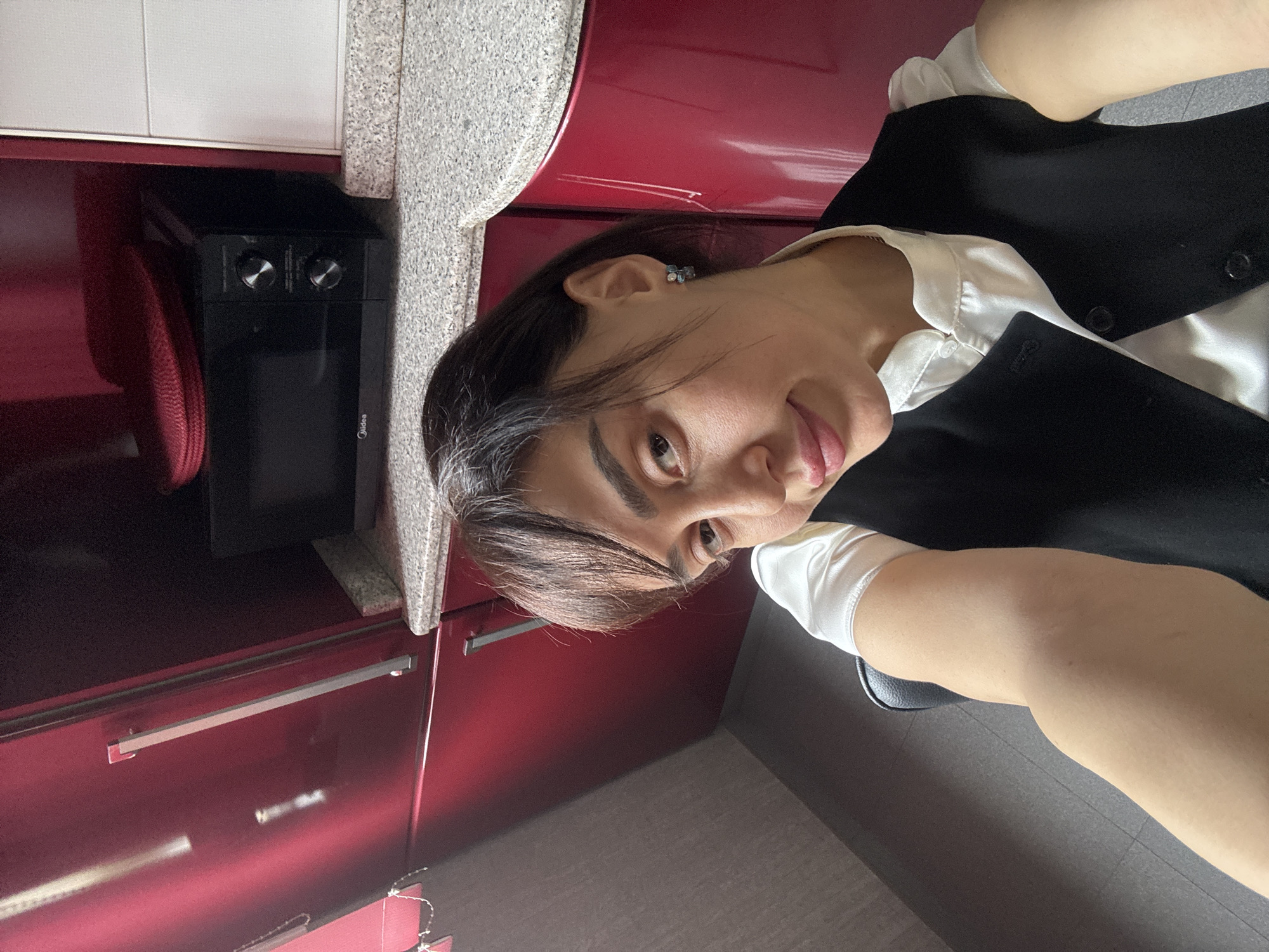 A tilted, indoor shot features a woman standing in what appears to be a kitchen. The kitchen has dark red cabinets with silver handles and a gray countertop. A black Midea microwave sits on the countertop, with what seems to be a red dish near it.
The woman is wearing a white collared shirt with a black vest over it. She has dark hair and is facing the camera with a slight smile. A small blue earring is visible. The background includes what appears to be a gray tile floor and a gray wall. The location is not discernible as Tbilisi, Georgia, without further contextual information.