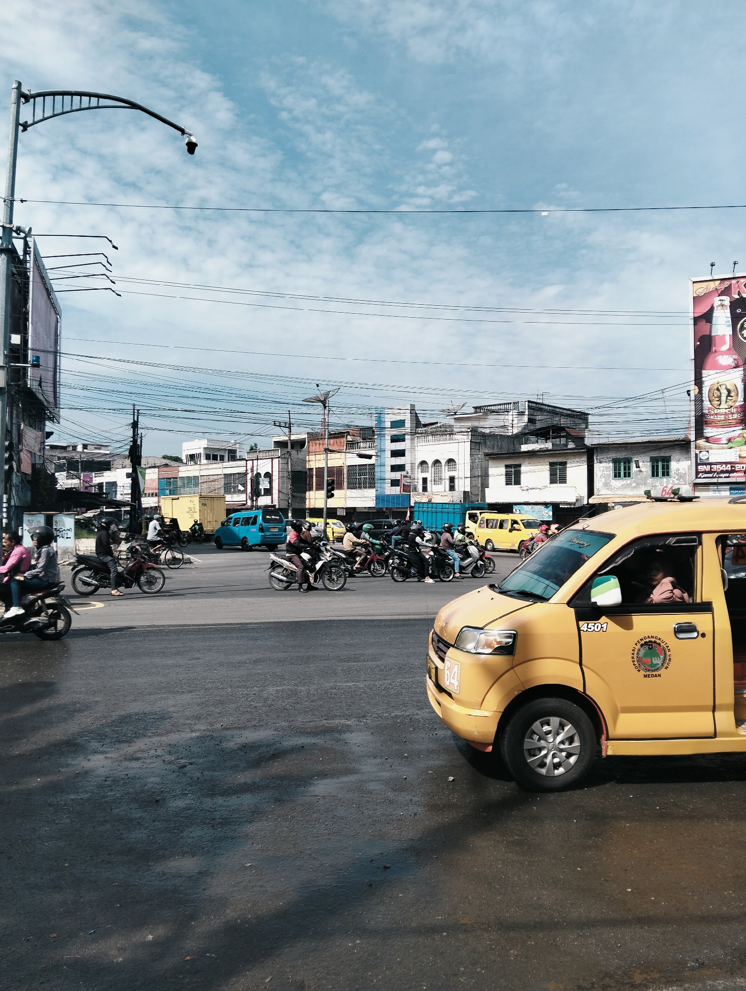 The image depicts a street intersection in the city of Medan, Indonesia, under a partly cloudy sky. In the foreground, a yellow taxi is prominently visible, positioned on the right side of the frame. The taxi has the number "4501" displayed on its side, along with a circular emblem bearing the words "KOPERASI PENGANGKUTAN UMUM" and "MEDAN". The taxi's door is slightly ajar, revealing a passenger inside. The road surface appears wet in patches, suggesting recent rain or a cleaning event, with shadows cast across it.
In the middle ground, a busy street scene unfolds. Numerous motorcycles, many carrying two riders, are navigating the intersection. Some motorcycles are moving, while others appear to be stopped or waiting. The riders are mostly wearing helmets. Further back, a blue van and a yellow minivan are visible. A yellow shipping container is also present on the side of the road.
The background is dominated by buildings of varying heights and architectural styles, typical of an urban environment. Power lines crisscross the scene, supported by poles. A street light with a security camera attached is visible on the left side of the image. A large billboard advertising a product, possibly a beverage, is situated on the far right. The overall impression is one of a bustling city intersection.