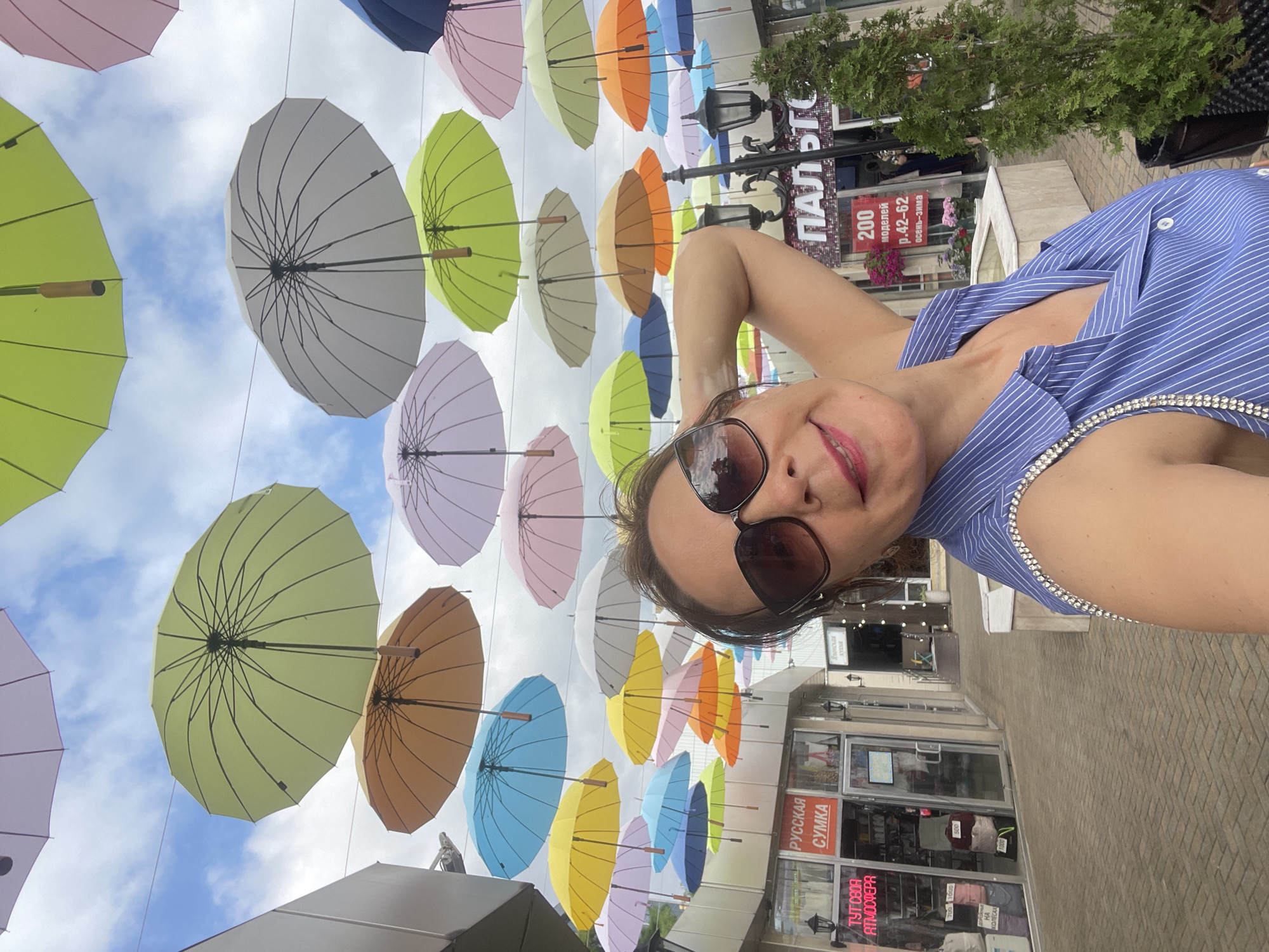 This is a vertical, outdoor selfie taken in an urban setting. A woman with dark hair, wearing sunglasses and a blue and white striped top adorned with rhinestones, smiles at the camera. She is positioned in the foreground, with her arm raised to take the photo.
Behind her, a vibrant display of colorful umbrellas is suspended overhead, creating a visually striking canopy. The umbrellas are in various shades of yellow, green, blue, pink, orange, and purple, against a cloudy sky. This installation appears to be part of a street decoration or an event.
In the background, there are storefronts with signage. One sign reads "РУССКАЯ СУМКА" (Russian Bag), and another indicates "ТУТ СВОЯ АТМОСФЕРА" (Your Own Atmosphere Here). Another sign visible mentions "ПАЛЬТО" (Coat) and "200 моделей р.42-62 осень-зима" (200 models sizes 42-62 autumn-winter). The ground is paved with brickwork. The overall impression is cheerful and festive, suggesting a daytime scene in a pedestrian area. The weather appears overcast. The context of Sochi, Russia, is provided.
