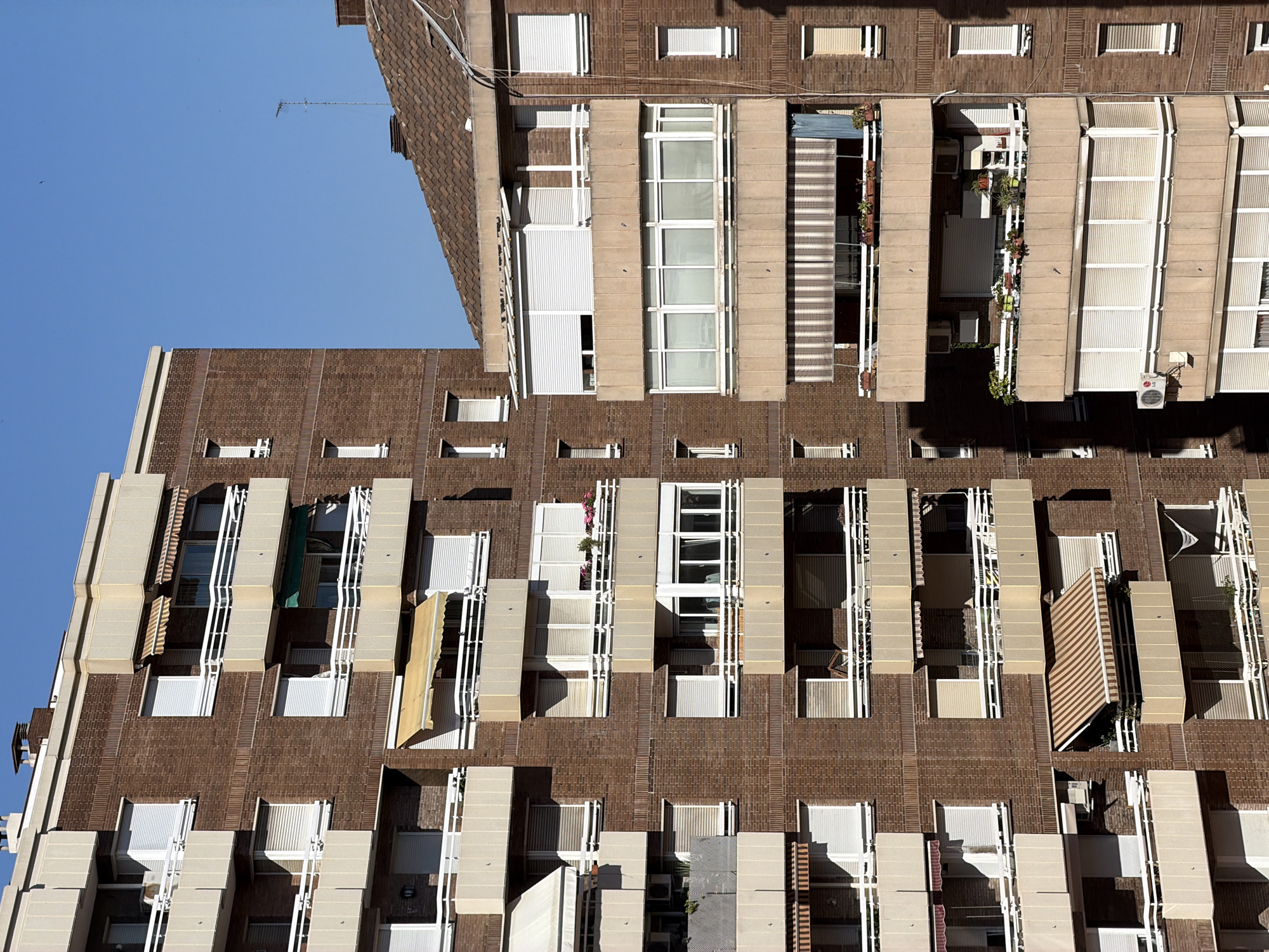 This media file showcases the exterior of a modern apartment building, likely located in Barcelona, Spain. The architectural style features a facade of reddish-brown brickwork interspersed with cream-colored concrete sections. The building has numerous windows and balconies, many equipped with white slatted shutters or awnings, suggesting a sunny climate.
The scene is characterized by an aerial perspective, looking down at an angle onto the building's facade. The clear blue sky in the background indicates fair weather. Several balconies are adorned with potted plants, adding touches of greenery. Visible objects include air conditioning units mounted on some of the balconies and a television antenna on a rooftop in the distance. There are no people visible in the frame, and the scene is devoid of any specific activity or event.
Notable details include the repetition of architectural elements like the vertical concrete supports and the patterned awnings, creating a sense of order and uniformity. The strong sunlight casts distinct shadows across the building's surfaces, suggesting it is daytime. No visible text is present in the image.