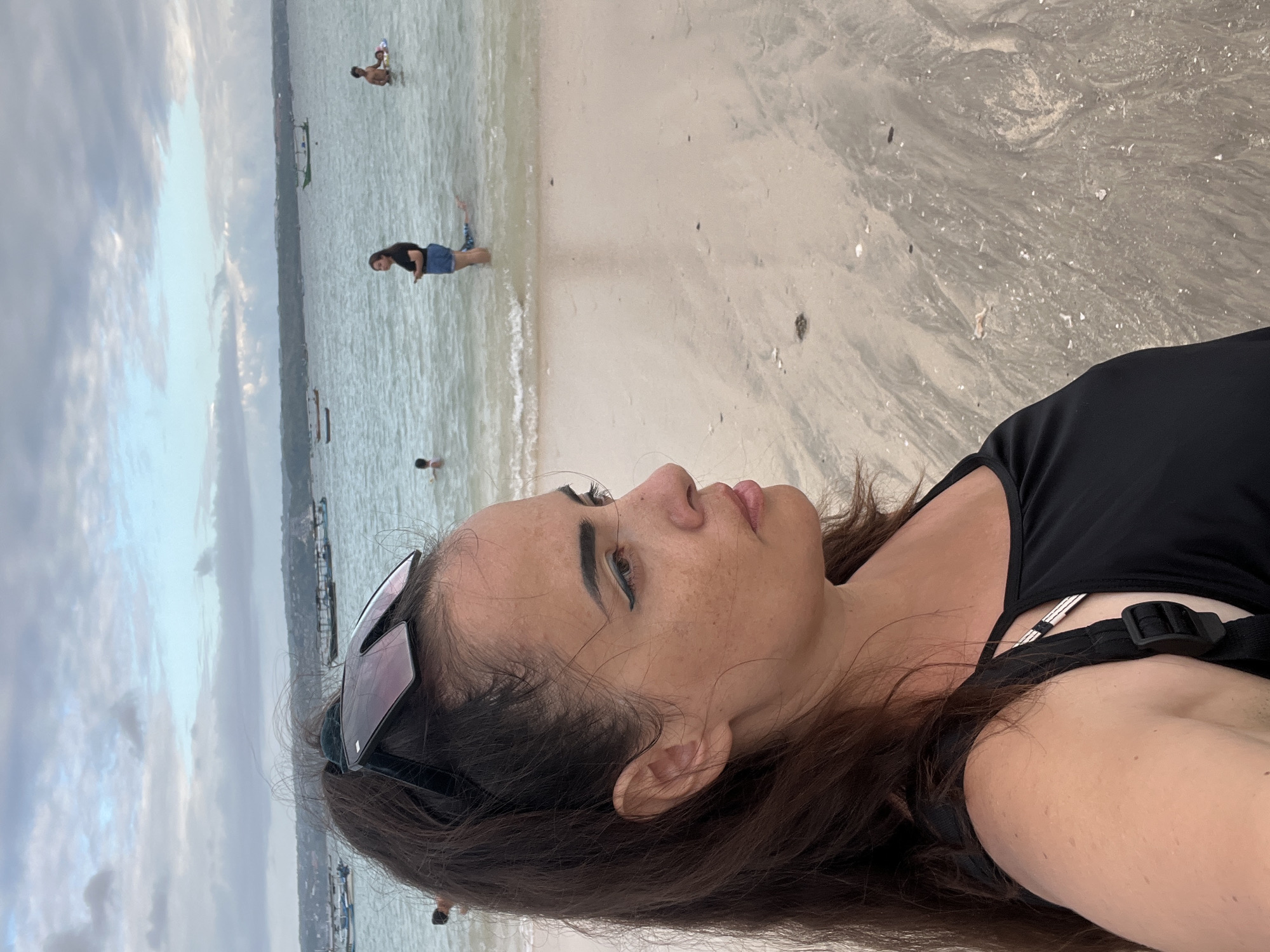This is a medium shot, taken from the chest up, of a woman standing on a sandy beach, with the ocean and a hazy horizon in the background. She is facing right in profile, her long, dark brown hair pulled up into a ponytail, with sunglasses perched on top of her head. She is wearing a black tank top. The beach sand is light brown with streaks of darker, wet sand, suggesting the tide has recently receded. Small shells and debris are scattered across the sand.
In the background, the ocean is a muted blue-grey, with gentle waves lapping the shore. Several small, traditional Indonesian fishing boats, known as jukungs, are visible dotted across the water, some closer to shore and others further out. A thin, green line of vegetation or land is visible along the horizon, with a cloudy sky overhead.
There are a few other people in the water in the distance. To the left, a child is partially visible in the water. Closer to the center, a woman in denim shorts and a black t-shirt stands in the shallow water, looking towards the left of the frame, holding a phone. To her right, a person, possibly a child, is partially submerged in the water, lying on their back. Further to the right, a man is visible in the water, standing and appearing to hold onto a yellow inflatable ring.
The scene depicts a casual day at the beach, likely in a coastal area near Denpasar, Indonesia, given the presence of jukung boats and the general atmosphere. The lighting suggests it may be early morning or late afternoon due to the soft, diffused light and the overcast sky.