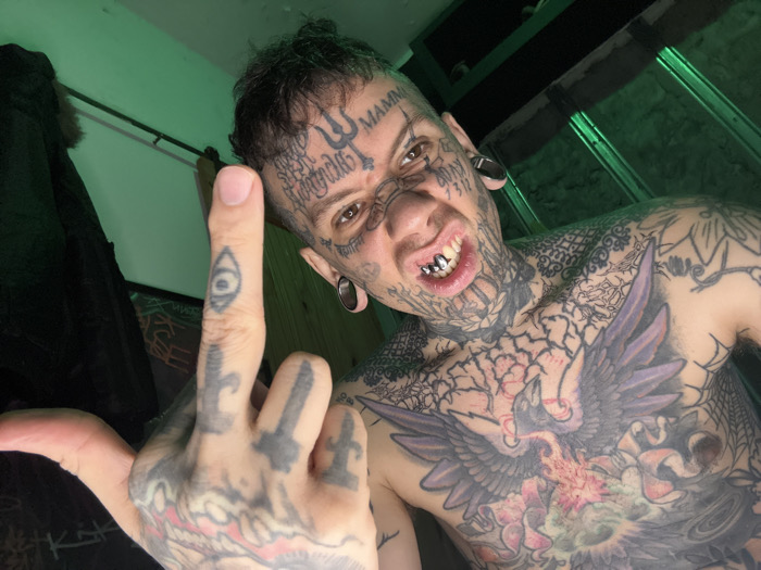 This image, captured indoors in Buenos Aires, Argentina, features a single individual, presumably male, extensively modified with tattoos and body jewelry. The subject's face, head, neck, chest, and visible left arm are covered in various tattoos, including intricate patterns, script, and symbols. On the forehead, a trident symbol, "MAMMA," and other text are visible, alongside numbers like "666" and "7312" near the right eye. The individual exhibits large ear gauges in both ears and a silver grill on the upper front teeth, exposed due to a grimacing or snarling facial expression. A prominent, multicolored tattoo depicting a large bird with outstretched wings, possibly a phoenix or eagle, surrounded by flame-like elements in purples, oranges, and greens, covers the subject's chest. The left hand is raised, with the middle finger extended towards the viewer, bearing an "all-seeing eye" tattoo, while an adjacent finger features an upside-down cross tattoo. The background is a dimly lit room, with a distinct green light source illuminating from the upper left. A dark fabric object hangs on the left side, and a horizontal bar is visible across the top of the background. A framed item with abstract patterns is partially discernible below the hanging fabric. The subject looks directly at the camera.