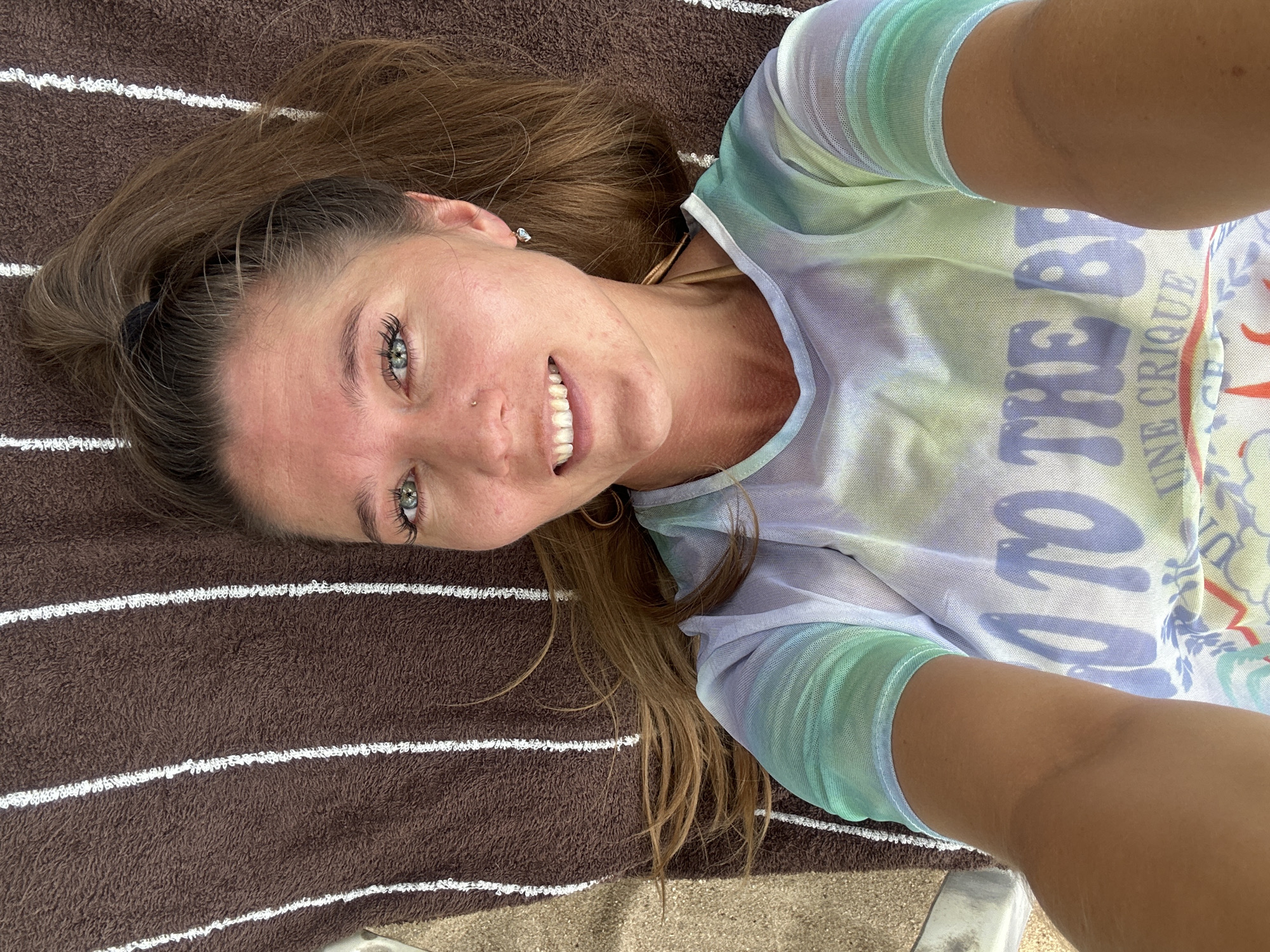 An overhead shot captures a woman lying on a brown towel with white stripes. She has long brown hair pulled up in a ponytail and is wearing a sheer, tie-dye t-shirt with the words "GO TO THE BEACH" visible on it. The woman has green eyes, a nose ring, and is smiling. The scene appears to be outdoors, possibly a beach or poolside area in Ko Samui District, Thailand.