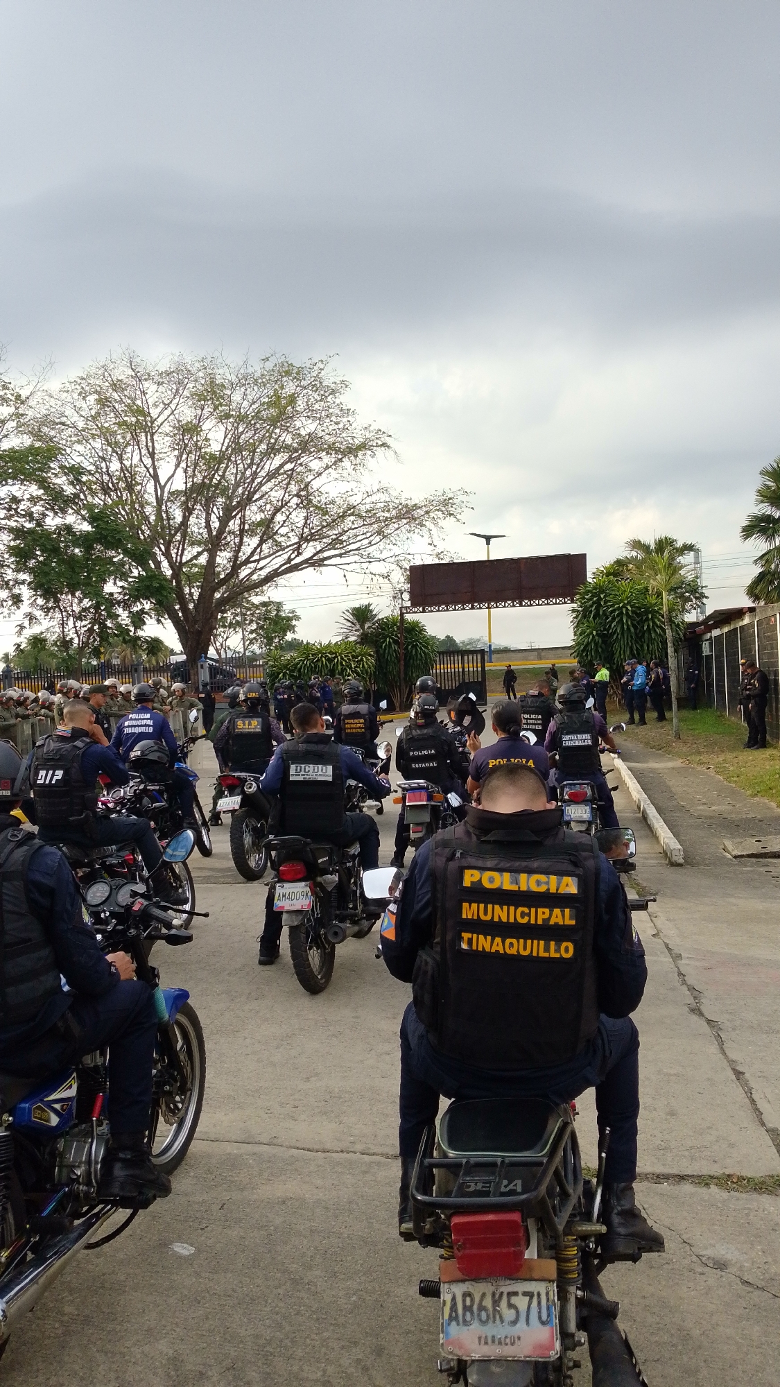 A medium-shot photo captures a gathering of numerous police officers on motorcycles, likely in Tinaquillo, Venezuela, given the visible text. The scene is set outdoors under an overcast sky, possibly during daytime. The officers are predominantly male, dressed in dark blue uniforms, tactical vests, and helmets. Most are seated on their motorcycles, which vary in model and age, suggesting a practical or utilitarian fleet. The officers appear to be assembled, possibly preparing for or returning from an operation, indicating a serious and professional atmosphere.
In the foreground and midground, officers on motorcycles face away from the viewer, their backs visible. Several tactical vests bear clear yellow lettering, such as "POLICIA MUNICIPAL TINAQUILLO," explicitly linking the location. Other vests display acronyms or names of different police units, including "S.I.P.", "DCDO," "POLICIA NACIONAL BOLIVARIANA," "POLICIA CONTRA REDES CRIMINALES," and "POLICIA ESTADAL," suggesting a multi-agency presence. License plates on the motorcycles are visible, with codes like "AB6K57U," "AM4D09K," and "AT2133G," some also featuring "YARACUY," a Venezuelan state.
The setting includes a paved surface, likely a road or a large parking area. To the left, a large, mature tree with sparse leaves stands, and palm trees are visible on the right. In the background, beyond a metal fence, there's what appears to be an empty billboard structure and other buildings, with more uniformed personnel gathered further in the distance. The overall impression is one of an organized police deployment or assembly in a somewhat rural or industrial outdoor environment.