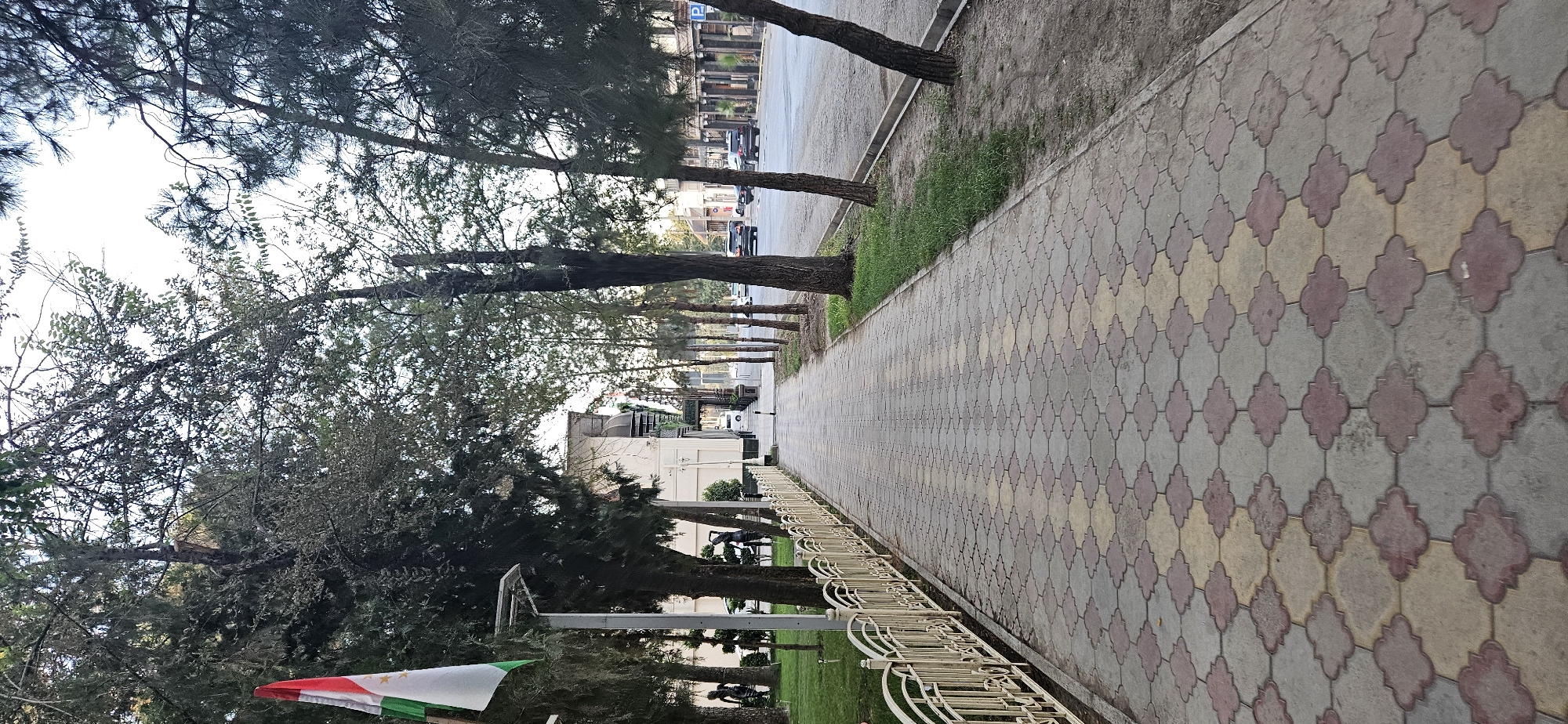 A paved sidewalk with a decorative pattern of light gray, pink, and yellow interlocking tiles runs along a tree-lined street in Dushanbe, Tajikistan. On the left side of the sidewalk, a cream-colored metal fence borders a grassy area with trees and a lamppost. A flag, appearing to be the flag of Tajikistan with its red, white, and green stripes and a crown, is attached to a pole near the flag. Across the street, there are buildings and parked cars. The street is paved and appears to be a road for vehicular traffic. The sky is overcast.