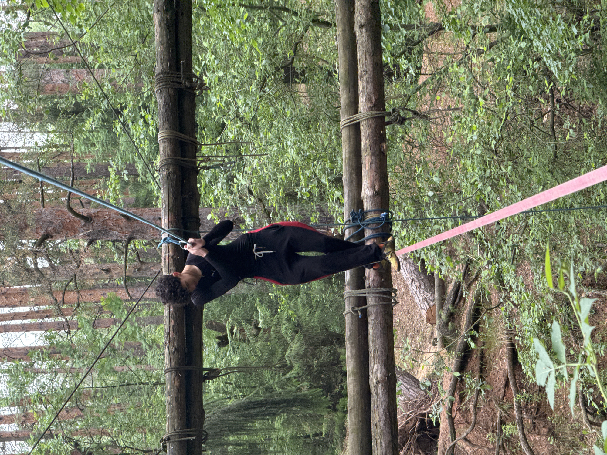 This image depicts an individual engaged in an outdoor ropes course or slacklining activity within a forest setting in Chaikovsky, Russia. A young person with dark, curly hair is seen actively balancing on a wide, pink-red slackline. They are wearing a black long-sleeved top with an indistinct white logo or symbol on the front, black trousers with red stripes running down the sides, and yellow or green athletic shoes.
The person is holding onto a parallel, thicker blue-green rope with both hands for stability, demonstrating focus and concentration. Both the slackline and the support rope are stretched horizontally between sturdy wooden poles or tree trunks, which are secured upright with additional ropes and knots. The background is dominated by lush green foliage from numerous trees, indicating a dense wooded area. The ground beneath the activity appears to be covered in natural forest debris. The lighting suggests an overcast daytime, providing soft, diffused light across the scene.