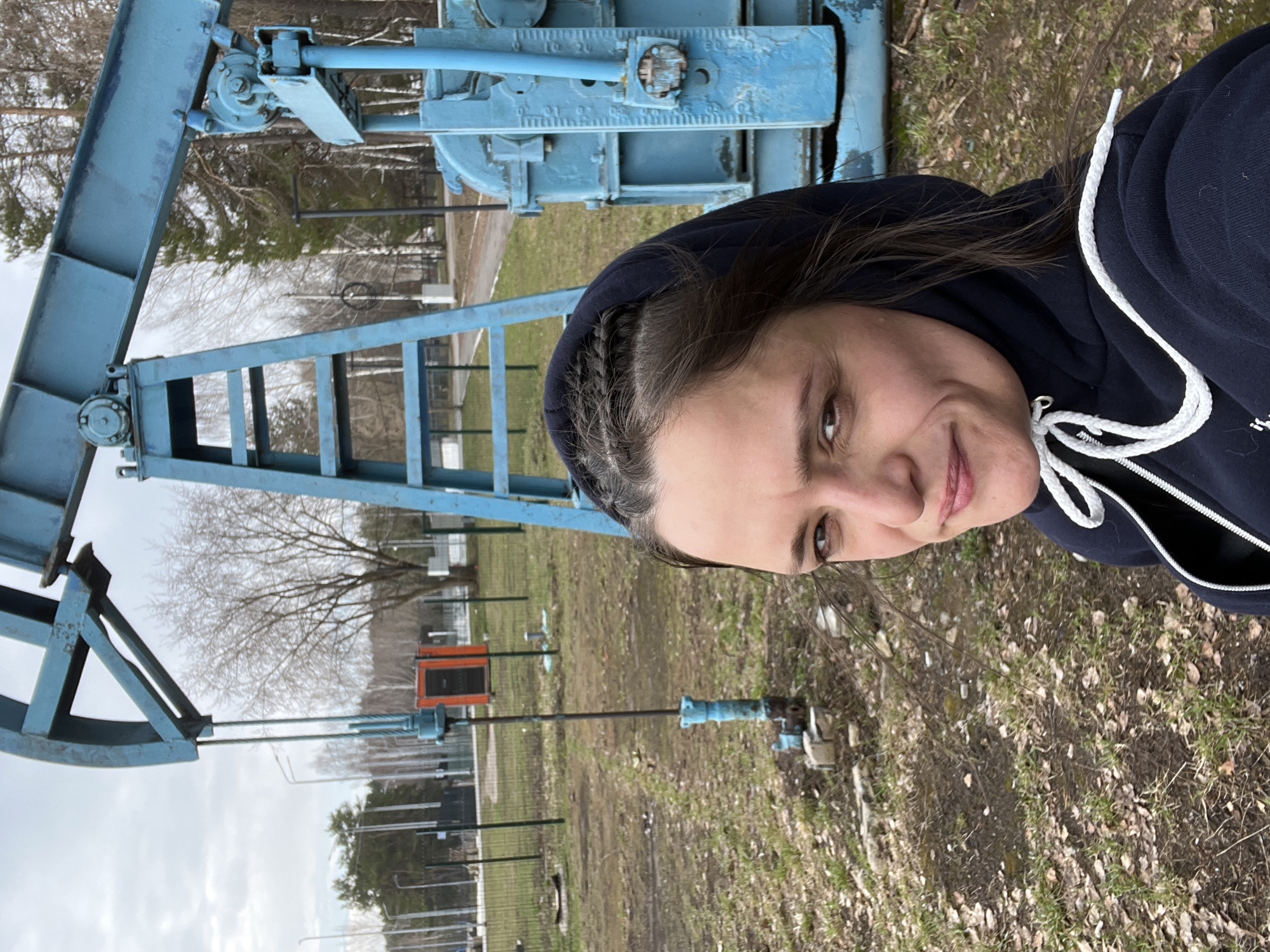 A woman in a dark blue hooded sweatshirt stands in front of an oil well pump jack (also known as a "nodding donkey") in Chaikovsky, Russia. The pump jack is painted light blue, and the surrounding ground is covered in dry grass, dead leaves, and patches of bare soil. In the background, a metal fence runs along the perimeter, with trees visible beyond. An orange and black rectangular structure is partially visible in the background as well, likely related to the pump jack system.