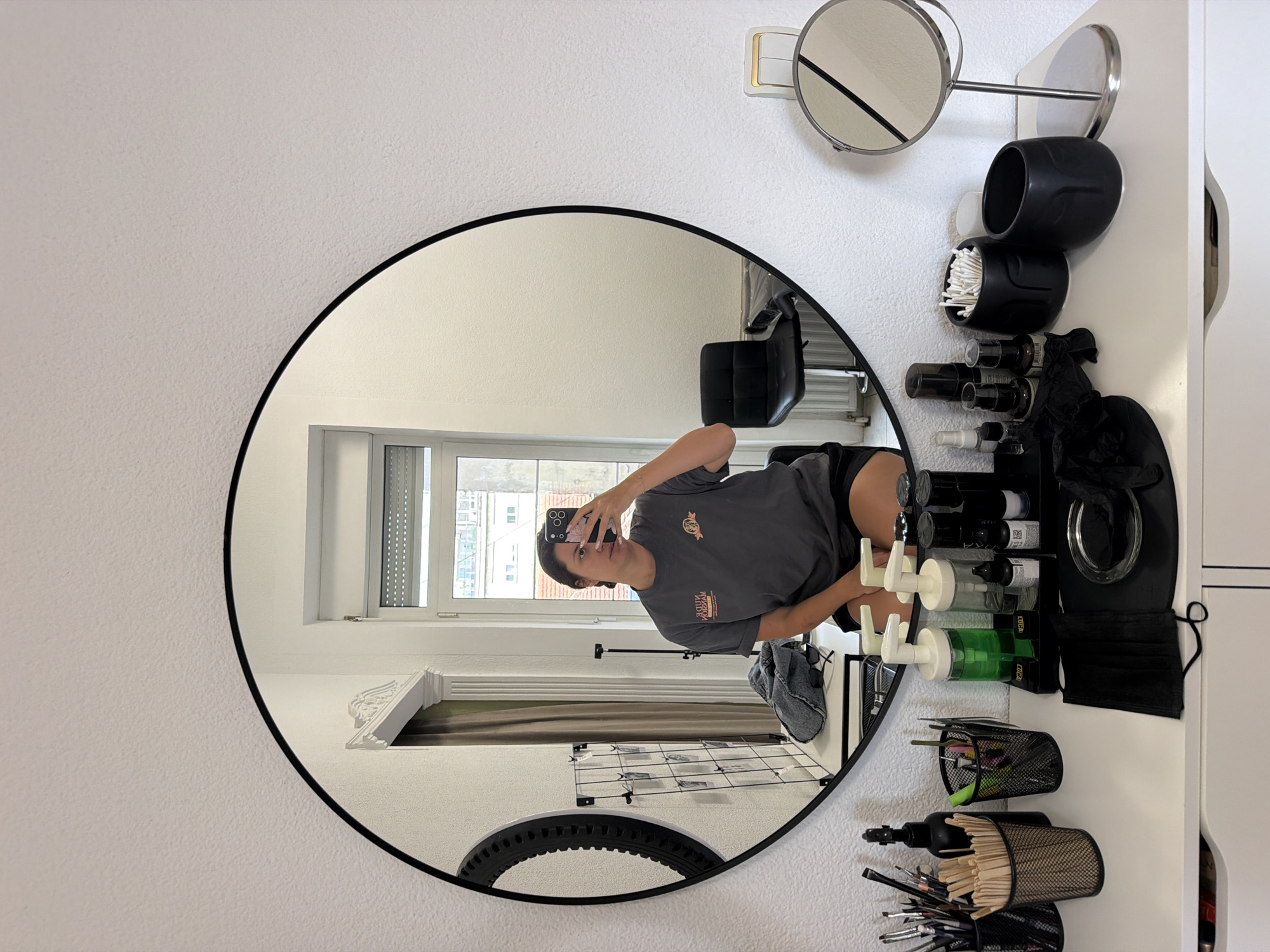 A woman is taking a mirror selfie in what appears to be a bedroom or studio in Belgrade, Serbia. She is wearing a grey t-shirt and black shorts, and is seated on a white dresser. The dresser is cluttered with various items, including makeup brushes, bottles of liquid, cotton swabs, and black gloves. A large circular mirror hangs on the wall behind her, reflecting the room. Also visible in the reflection is a window, a black chair, and a radiator.