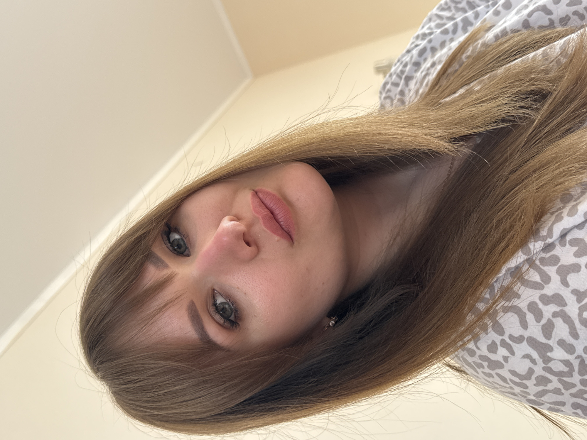 A photograph shows a woman with long brown hair, fair skin, and gray eyes looking down at the camera. She has bangs that partially cover her forehead. She is wearing a white shirt with a gray animal print. The background consists of a plain, light-colored wall, suggesting an indoor setting. The image has a tilted perspective, taken from above. There is no evidence to indicate the location is Yevpatoriya, Ukraine.