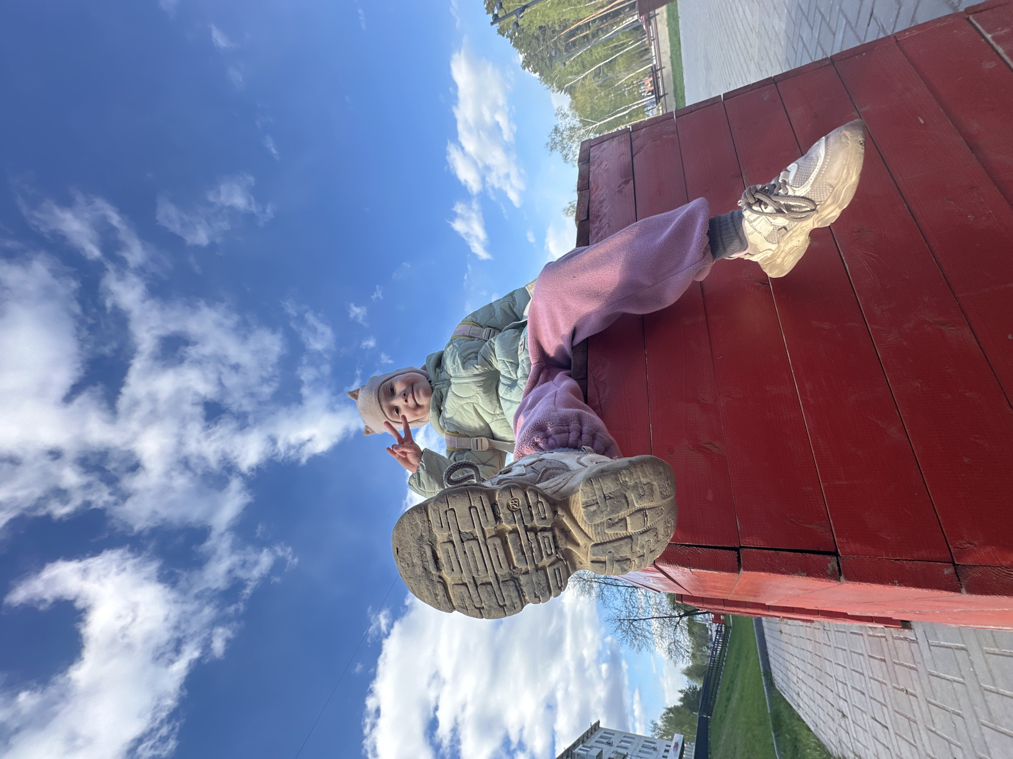 A girl sits atop a red wooden structure. She wears a light green jacket, pink pants, a light-colored hat with pointed ears, and white sneakers. She holds up a peace sign. The soles of her shoes are visible, with one appearing significantly closer to the camera than the other. The background consists of a blue sky with scattered white clouds. Trees, a sidewalk, a fence, a building and grass are partially visible in the distance. The image was taken from the location in the city of Chaikovsky, Russia.