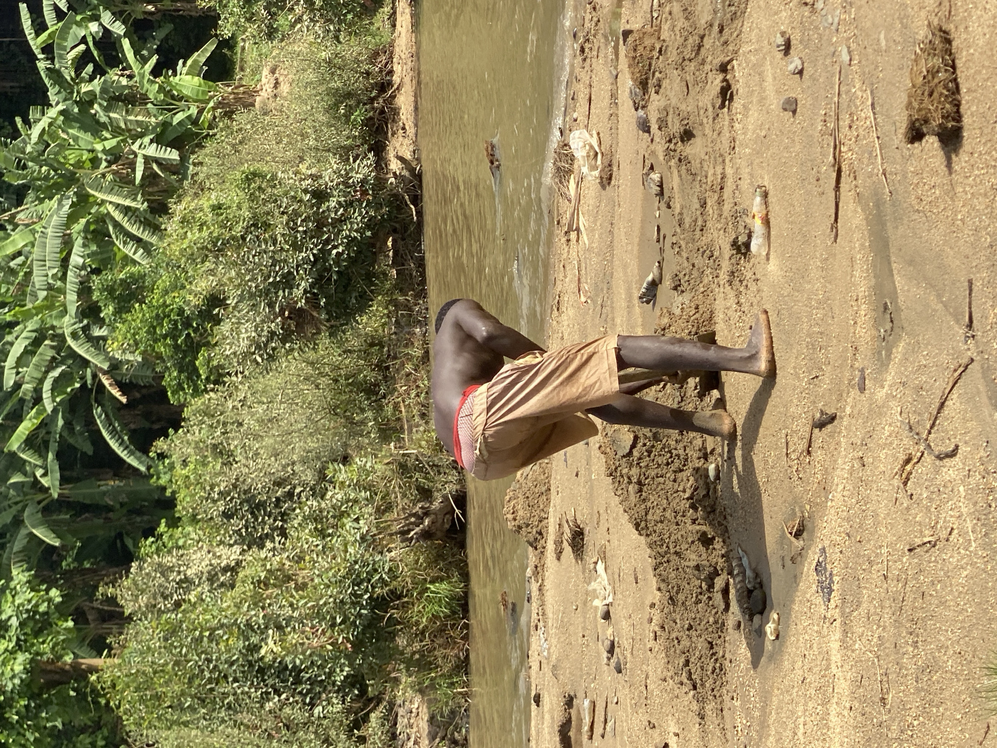 The image, viewed upright, depicts a natural outdoor scene at a riverbank in Maigizo, Nigeria, under bright daylight. A person with dark skin, wearing light brown shorts and a red-banded underwear band, is prominently featured in the right foreground. They are bent over at the waist, facing a body of water, with their bare feet on the sandy and muddy bank. Their posture suggests an activity involving interaction with the water or the ground, such as washing or collecting.
The immediate bank on which the person stands is sandy and uneven, with small mounds of dirt and scattered debris, including a small plastic bottle, sticks, and pebbles. In the middle ground, a murky, brown-colored body of water flows, exhibiting ripples on its surface. Several pieces of light-colored litter, including a plastic bag, are visible floating or partially submerged in the water, particularly near the far bank.
The far bank, in the left background, is covered with dense, lush green vegetation, featuring numerous large, broad leaves resembling those of banana plants or similar tropical flora. The strong sunlight casts distinct shadows, indicating a clear, sunny day. No text is visible in the image. The setting suggests a rural or undeveloped area.