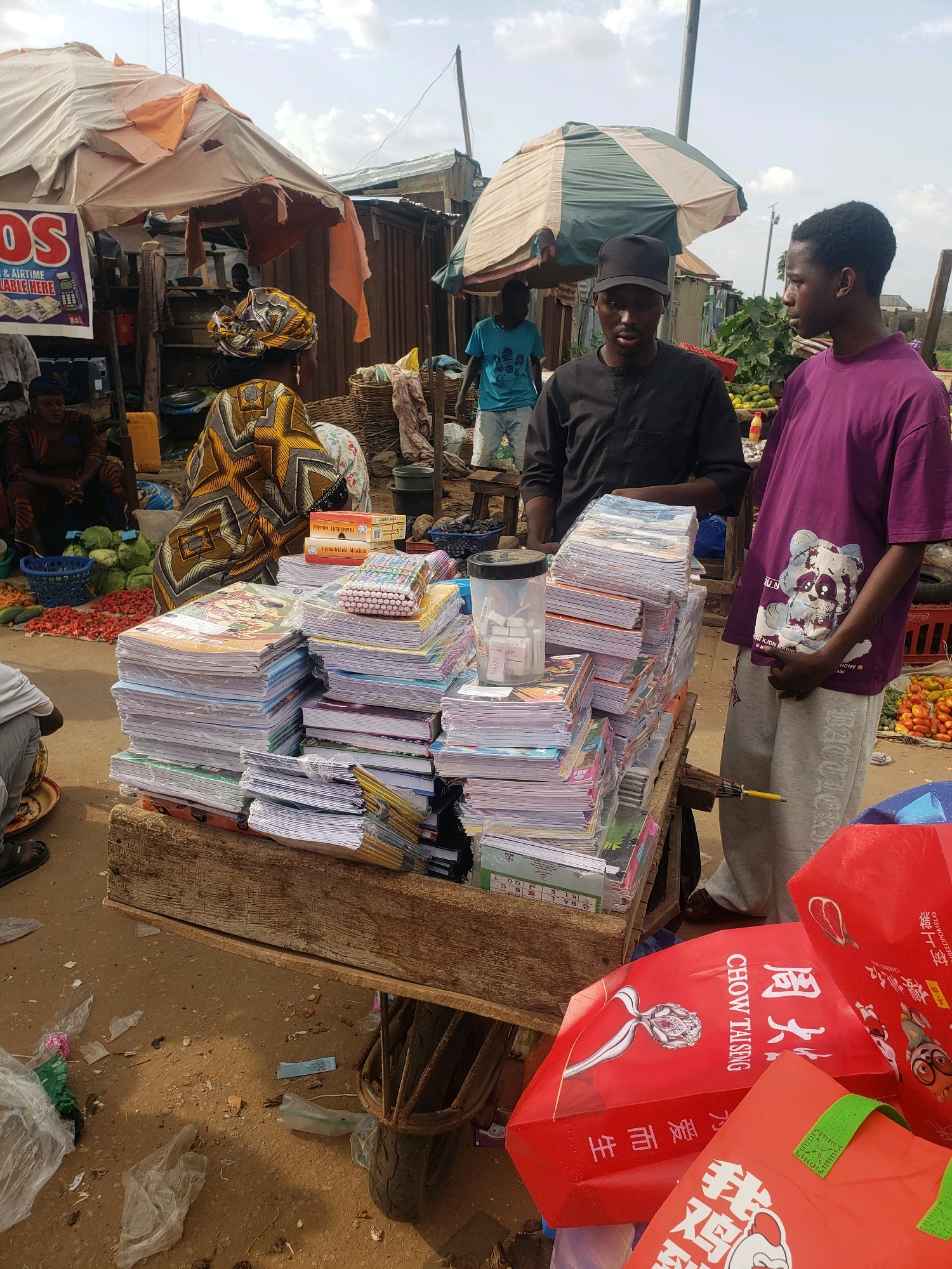 In Sabon Tasha, Nigeria, a market scene unfolds under a cloudy sky. The foreground is dominated by a wooden cart overflowing with stacks of notebooks and stationery, some wrapped in plastic. A partially visible person wearing a white shirt and grey pants stands to the right, looking towards the cart. To the left of the cart, a man in a black cap and black traditional attire engages in conversation with the person in white. In the background, several market stalls with makeshift roofs are visible, some displaying goods like fruits and vegetables. The ground is dusty and littered with plastic bags and debris. The overall atmosphere suggests a bustling, outdoor market environment.