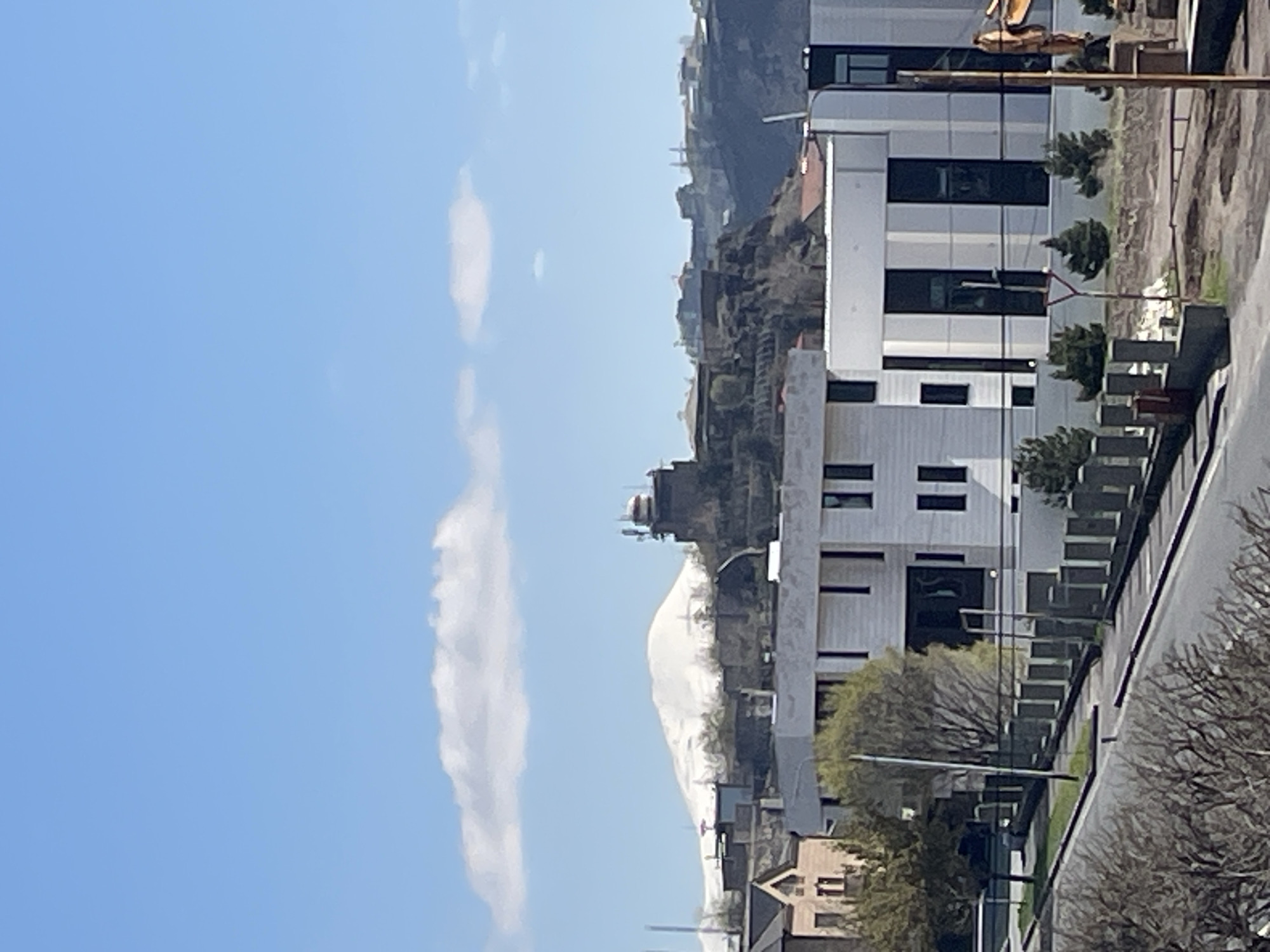 The image depicts a cityscape under a blue sky with elongated white clouds. A modern white building with multiple dark-framed windows stands prominently in the foreground. Adjacent to it is a street scene featuring trees and an urban landscape. In the background, a mountain or hill is visible with buildings perched on its slopes. In the distance, a snow-capped mountain. The location is Yerevan, Armenia.