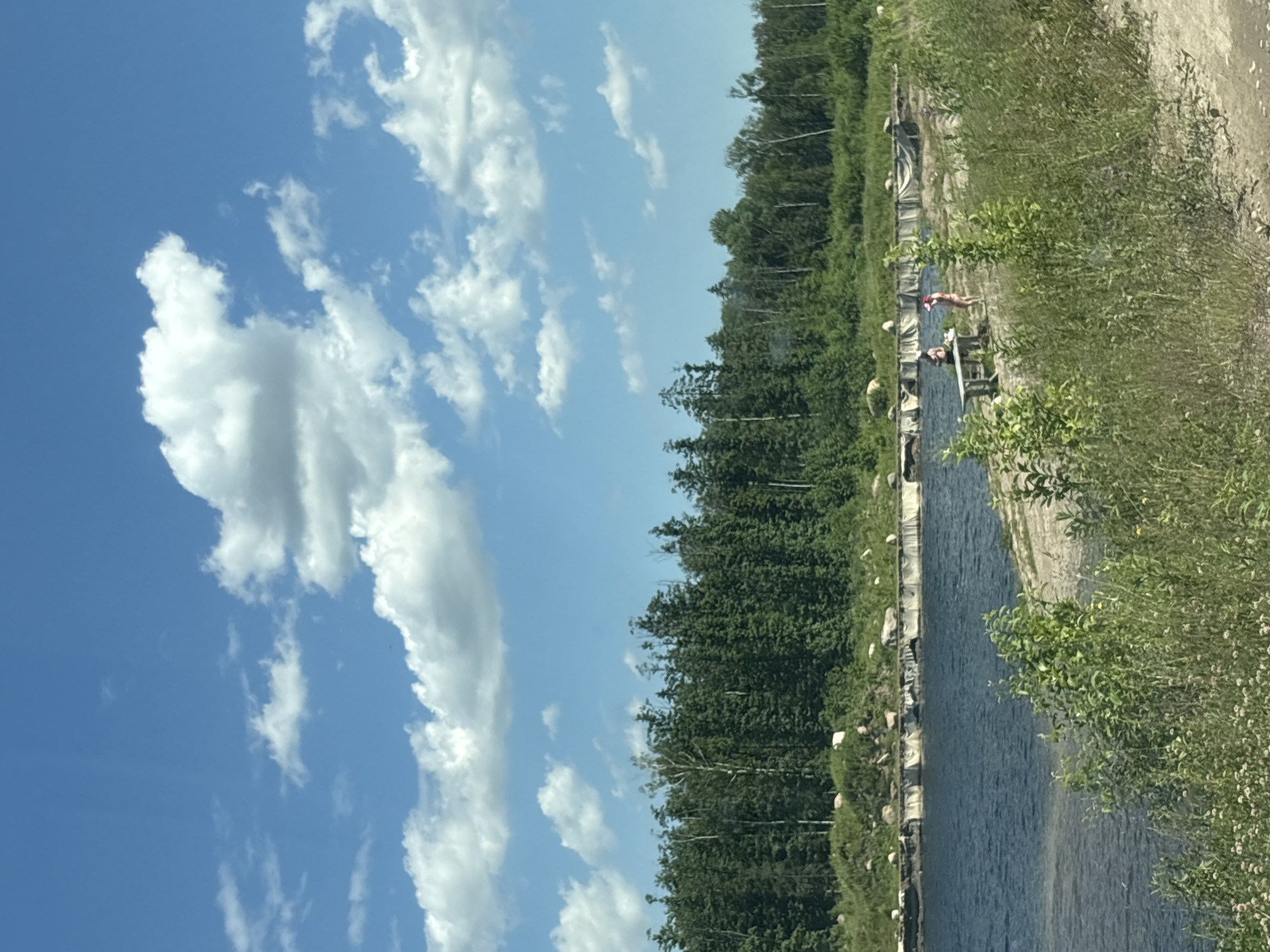 The image depicts a serene natural landscape with a body of water in the foreground, bordered by a sandy bank and lush vegetation. In the middle ground, a line of trees forms a dense forest against a bright blue sky dotted with white, puffy clouds. Two individuals are visible on the sandy bank near the water's edge. One person is standing, and the other is seated at a wooden picnic table. The setting appears to be a recreational area or a natural spot for relaxation, possibly a lake or a quarry.