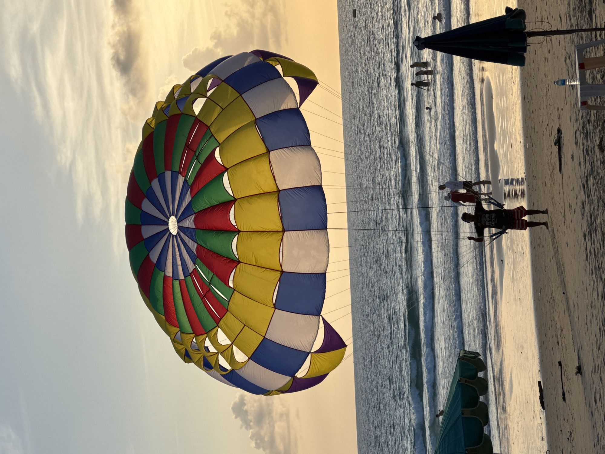 At sunset on Karon Beach in Karon, Thailand, a large, colorful parachute used for parasailing dominates the sky. The parachute is a vibrant mix of red, green, blue, yellow, and white segments, with its canopy partially inflated and suspended by rigging lines. Below the parachute, a person stands on the wet sand near the water's edge, holding onto the parachute's control lines. They are wearing striped shorts and a dark t-shirt. In the background, the calm ocean has gentle waves rolling onto the shore. Several people are visible in the shallow water, some wading and others appearing to swim. The beach itself is sandy, with discarded debris like driftwood scattered across it. To the left, a large green beach umbrella is partially visible, and to the right, a dark blue umbrella stands on the sand, next to a white plastic table with a water bottle. The sky is a soft orange and yellow, with scattered clouds illuminated by the setting sun.
