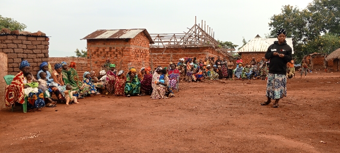 A group of people, mainly women and children, is gathered in a rural setting. They are seated or standing in a semi-circle, listening to a woman who is standing and gesturing towards them. Behind the group are several brick buildings, some with corrugated iron roofs and one under construction with exposed wooden beams. The ground is red earth, and there are trees and other buildings in the background. The sky is overcast.