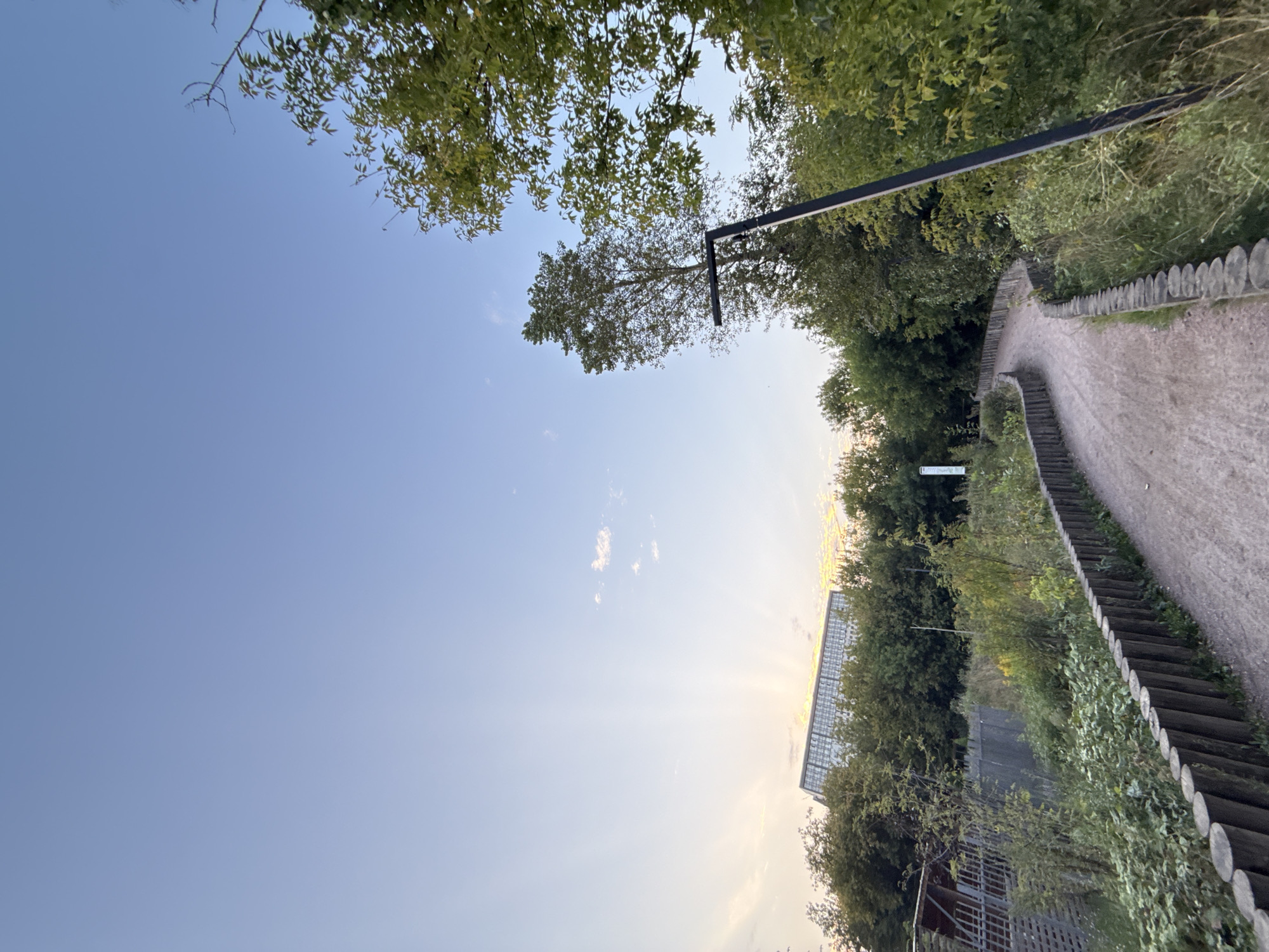 This is a low-angle, eye-level shot of a winding dirt path in a park-like setting at dusk. The path is bordered on the left by a low wooden fence made of logs and on the right by lush green vegetation. A tall, dark lamppost stands on the right side of the path. In the background, behind a dense line of trees, a modern glass-fronted building with a distinctive geometric shape is visible, bathed in the orange glow of the setting sun. The sky is a clear, pale blue with a few wisps of clouds near the horizon, and sun rays emanate from behind the building. The scene suggests a peaceful, urban natural space. The building in the background is likely the Moscow Planetarium, judging by its unique architecture and location relative to a park.