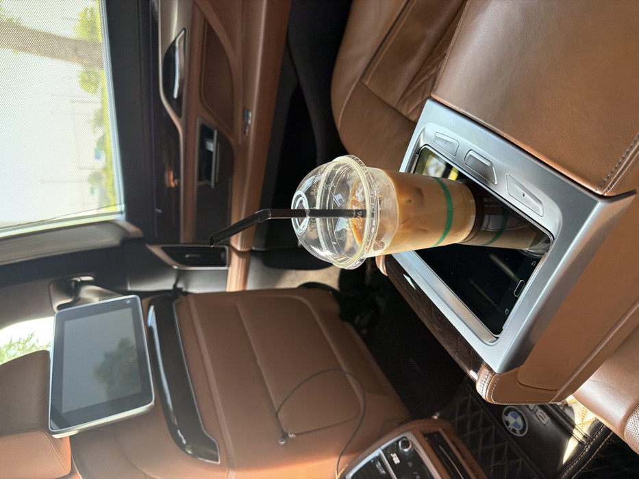 The image depicts the interior of a luxury BMW vehicle, specifically the rear passenger compartment. A clear plastic cup filled with iced coffee, complete with a black straw and a green band near its base, rests on a rear center console. This console incorporates a dark-screened electronic device, possibly a tablet or control panel, flanked by silver physical buttons and brown leather upholstery. Brown leather seats are prominent throughout the cabin, complemented by polished wooden trim and silver accents. A rear-seat entertainment screen is mounted on the back of the front passenger seat headrest, from which a wired earphone cable hangs. A black quilted floor mat bearing the BMW logo is visible in the footwell. Through the partially shaded rear window, green foliage and a distant road or infrastructure are visible under daylight conditions. No individuals are present. The scene is set within a vehicle in the city of Dubai, United Arab Emirates.