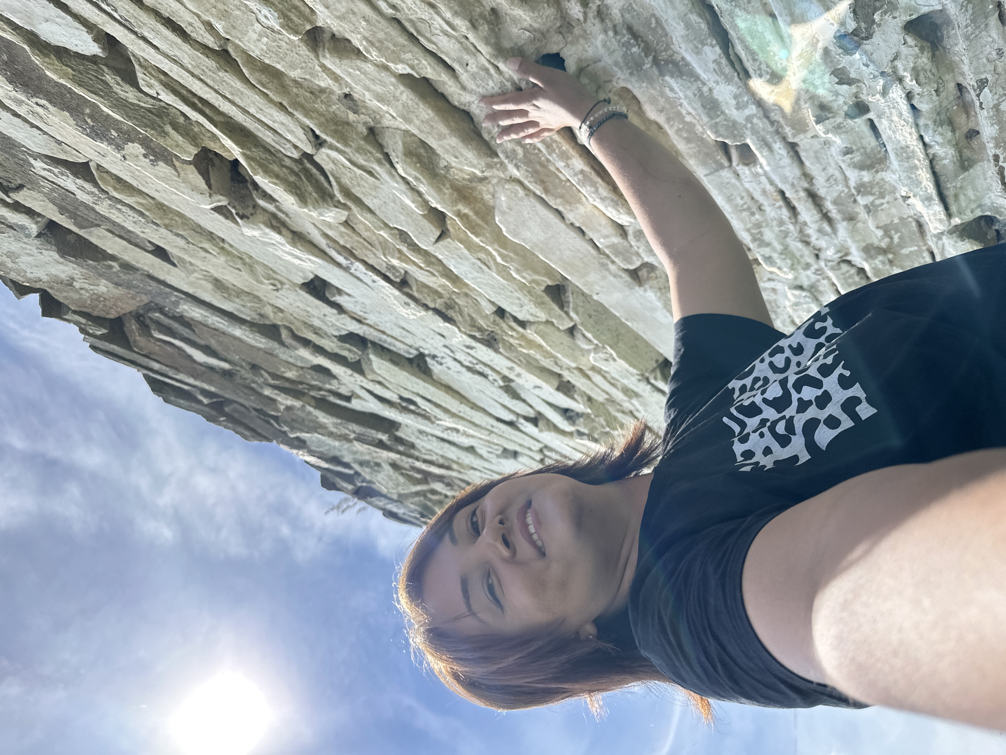 A low-angle, medium shot captures a woman in a black t-shirt with a white, leopard-print design on the right side of her chest. She is positioned in the foreground, looking upwards towards the right side of the frame with a smile. Her left arm is extended, and her hand is touching a rough, light-colored stone wall that occupies the majority of the right side of the image. The wall is constructed of irregular, stacked stones, suggesting an outdoor, possibly natural setting. The sky, a bright blue with scattered white clouds, is visible in the upper left quadrant, with the sun shining brightly. The woman has brown hair, and wears bracelets on her left wrist. The overall scene suggests an outdoor activity in a sunny environment.