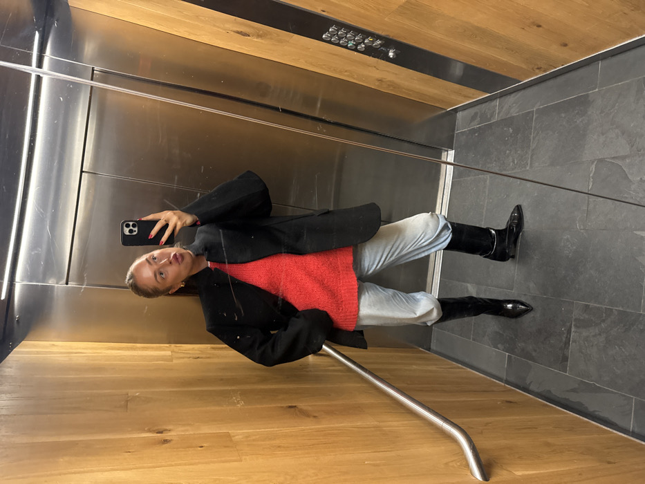 The image depicts a female individual taking a self-portrait inside an elevator, located in the city of London, United Kingdom. The subject, positioned centrally, holds a dark-colored smartphone in her left hand, which displays a camera interface. Her fingernails are painted red, and a ring is visible on her left hand. Her right hand is placed inside the pocket of her black blazer. She wears a bright red, possibly knitted, long-sleeved top beneath the blazer, light-colored, loose-fitting trousers, and black, glossy, pointed-toe boots. Her hair is pulled back, and she has visible hoop earrings. Her lips are pursed. The elevator interior features reflective metallic doors on the opposing wall, displaying visible scratches and reflecting an elevator control panel with multiple silver buttons. The side walls are constructed with vertical wooden panels, and a curved, silver-toned metal handrail extends along the left wall. The floor is composed of dark gray or black tiles with visible grout lines.
