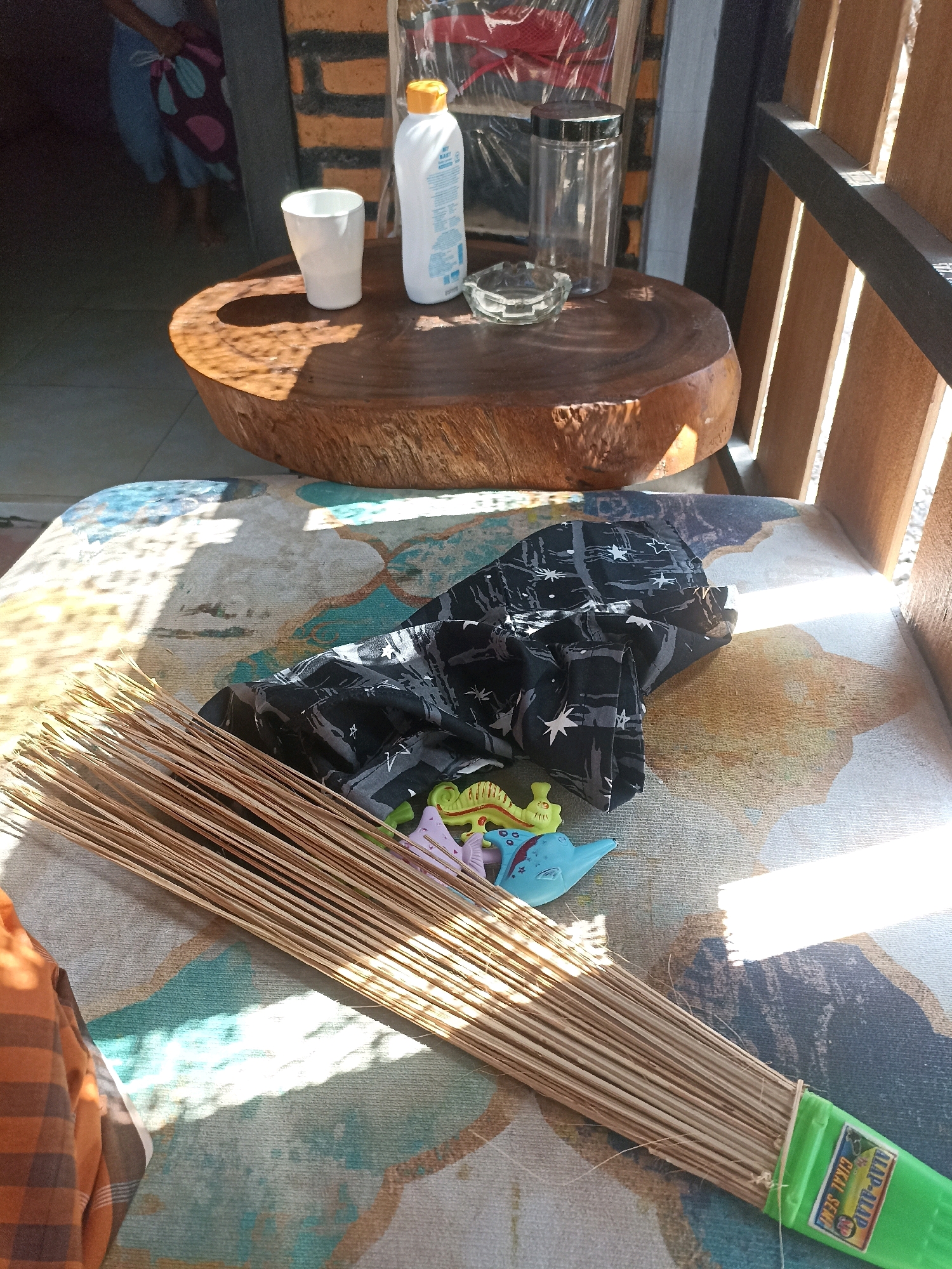 An outdoor scene, likely a patio or veranda in Mojopanggung, Indonesia, features a variety of objects placed on a cushioned seat. A large, round wooden table with a rough-hewn edge is positioned behind the seat. On the table, a white ceramic cup, a bottle of white liquid with a yellow cap, an ashtray, and a clear glass jar with a black lid are visible.
On the cushioned seat, a bundle of dry, thin wooden sticks, resembling a broom, lies diagonally across the foreground. The bundle is secured with a green plastic band, which has a label attached. In front of the broom, a black fabric with a white star pattern is folded. Several small, colorful plastic toys, including what appears to be a blue dinosaur and a yellow chameleon, are scattered near the broom.
The background shows a wall constructed of orange bricks with dark mortar, and part of a wooden fence or partition with vertical slats. Sunlight casts distinct shadows across the scene, indicating a bright, sunny day. In the far left background, a person's legs and lower torso are visible, wearing blue pants and holding a patterned bag. The overall impression is one of casual, outdoor living space.