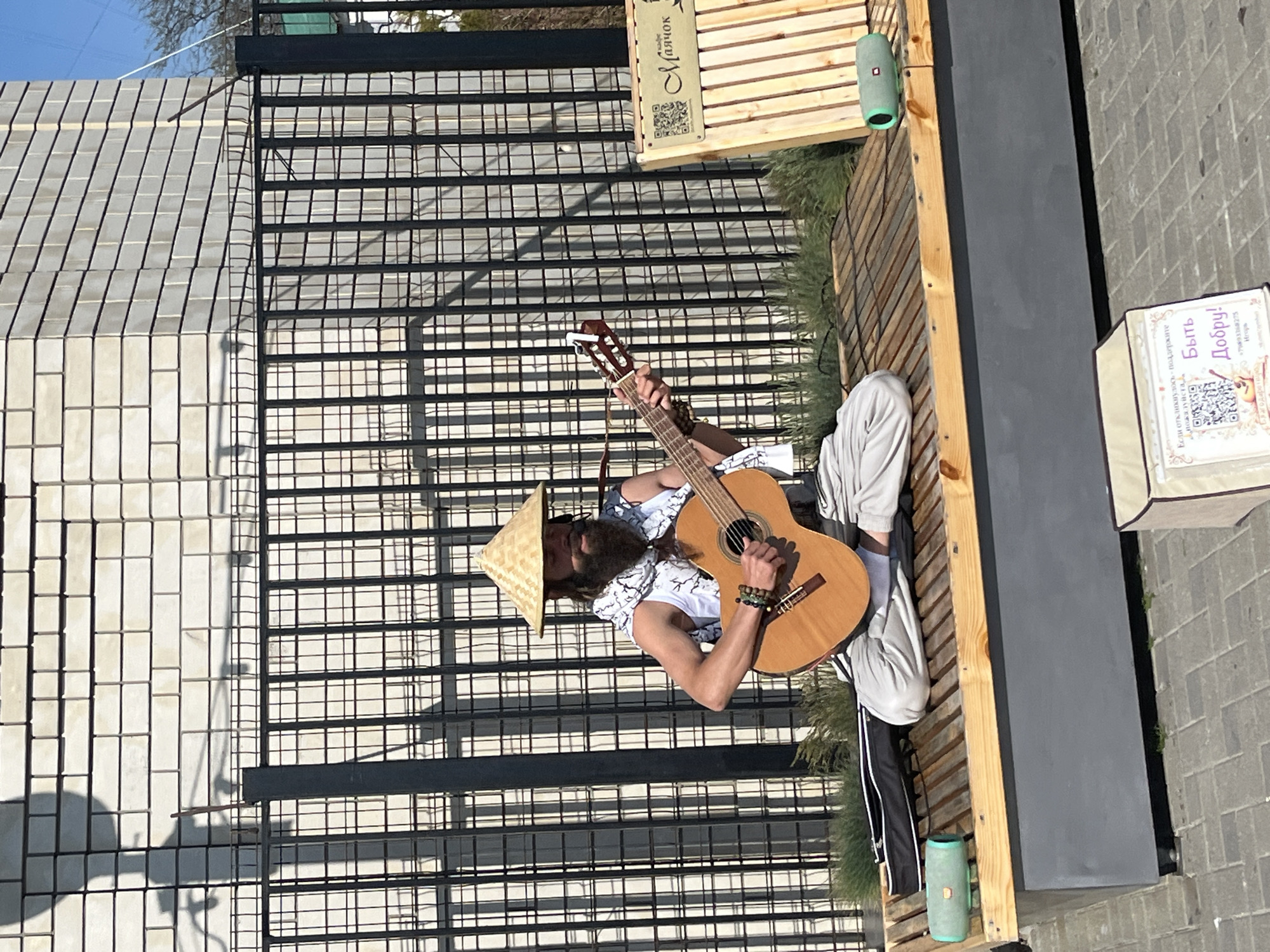 A street performer playing a guitar in Anapa, Russia, sits cross-legged on a wooden bench near a black wrought iron fence. The performer wears a conical Asian-style hat, a white printed shirt, and light-colored pants. The bench is adjacent to a dark grey rectangular column. A sign and a speaker are also on the bench. The ground is paved. A building of light-colored bricks is visible behind the fence.