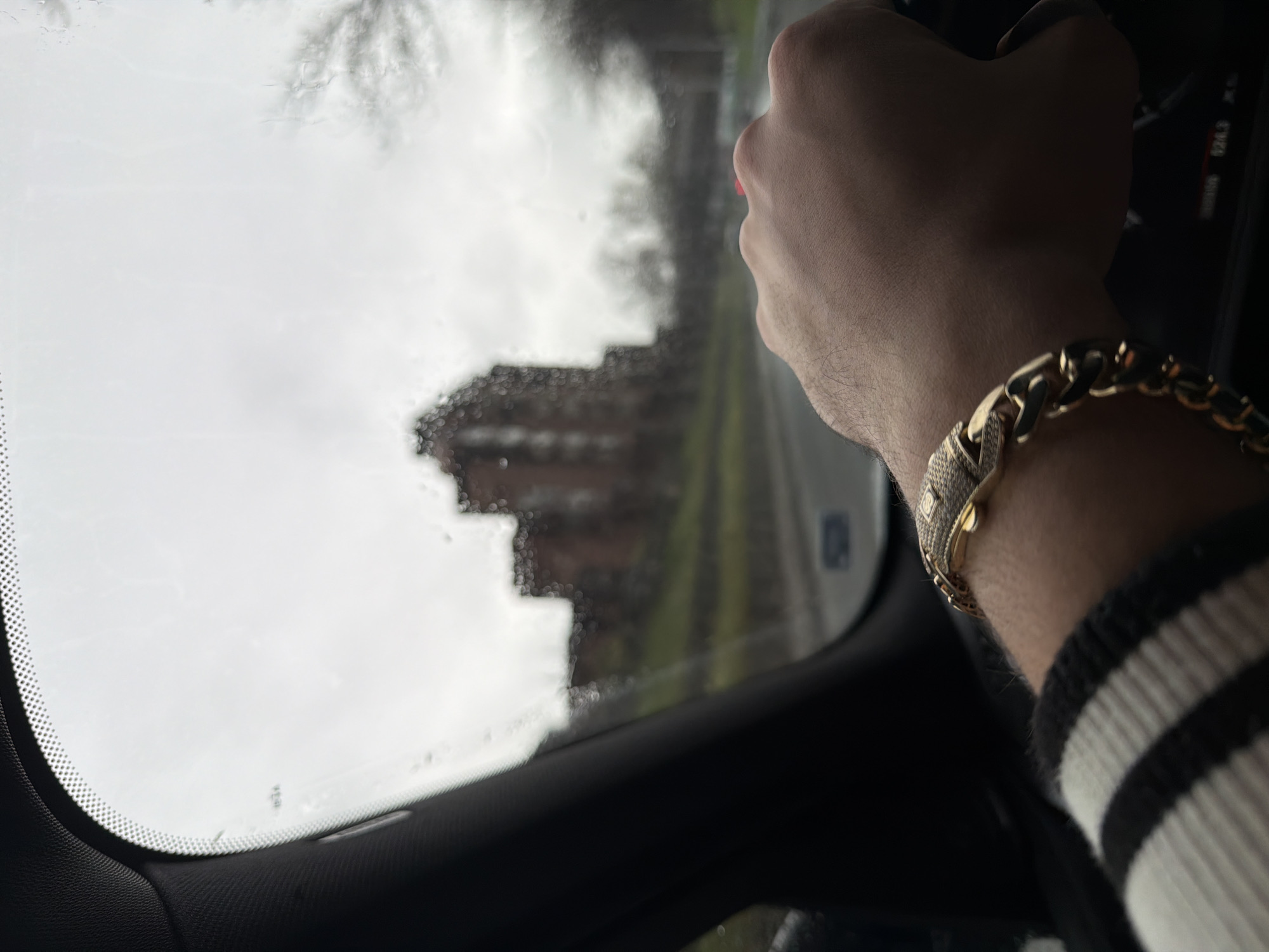The image shows the interior of a car, specifically the driver's side. A person's hand and wrist are visible, adorned with a thick gold chain bracelet that appears to have embedded stones. The person's hand is positioned near the steering wheel, suggesting they are driving. Through the windshield, it's raining and the view is somewhat obscured, but a large building is discernible, indicating an urban environment. The location is Plovdiv, Bulgaria.