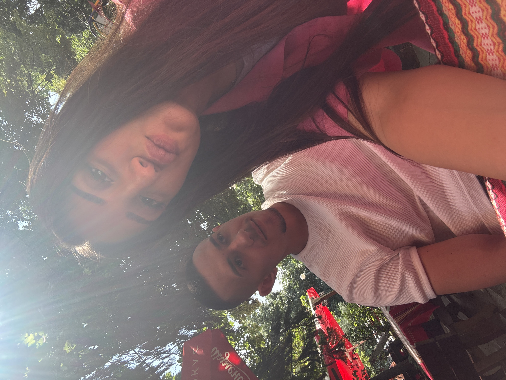 This rotated image captures a selfie of two individuals, a woman and a man, enjoying an outdoor setting in Plovdiv, Bulgaria. The photo appears to have been taken from an elevated perspective, possibly with a phone held above them.
The woman, positioned in the upper left portion of the frame, has long, dark hair and is wearing a light pink top. Her face, including her eyes and lips, is partially visible through a bright lens flare caused by the sun. Adjacent to her, a colorful, vertically striped fabric with geometric patterns, possibly a bag or a blanket, adds a vibrant detail.
The man is seen in the lower right, looking towards the camera. He has short, dark hair and a neatly trimmed beard or stubble. He is wearing a white, textured short-sleeved polo shirt. Both individuals maintain a neutral and relaxed expression.
The scene is set outdoors during daylight hours, with ample sunlight filtering through dense green foliage and trees, creating bright highlights and some lens flare. In the background, several large red umbrellas, likely belonging to a cafe or restaurant, are visible. Faint, inverted text can be discerned on one of the red umbrellas, possibly including words like "OCHOBA" or "VERMOUTH," suggesting a commercial establishment. The overall atmosphere is bright and casual, indicating a sunny day.