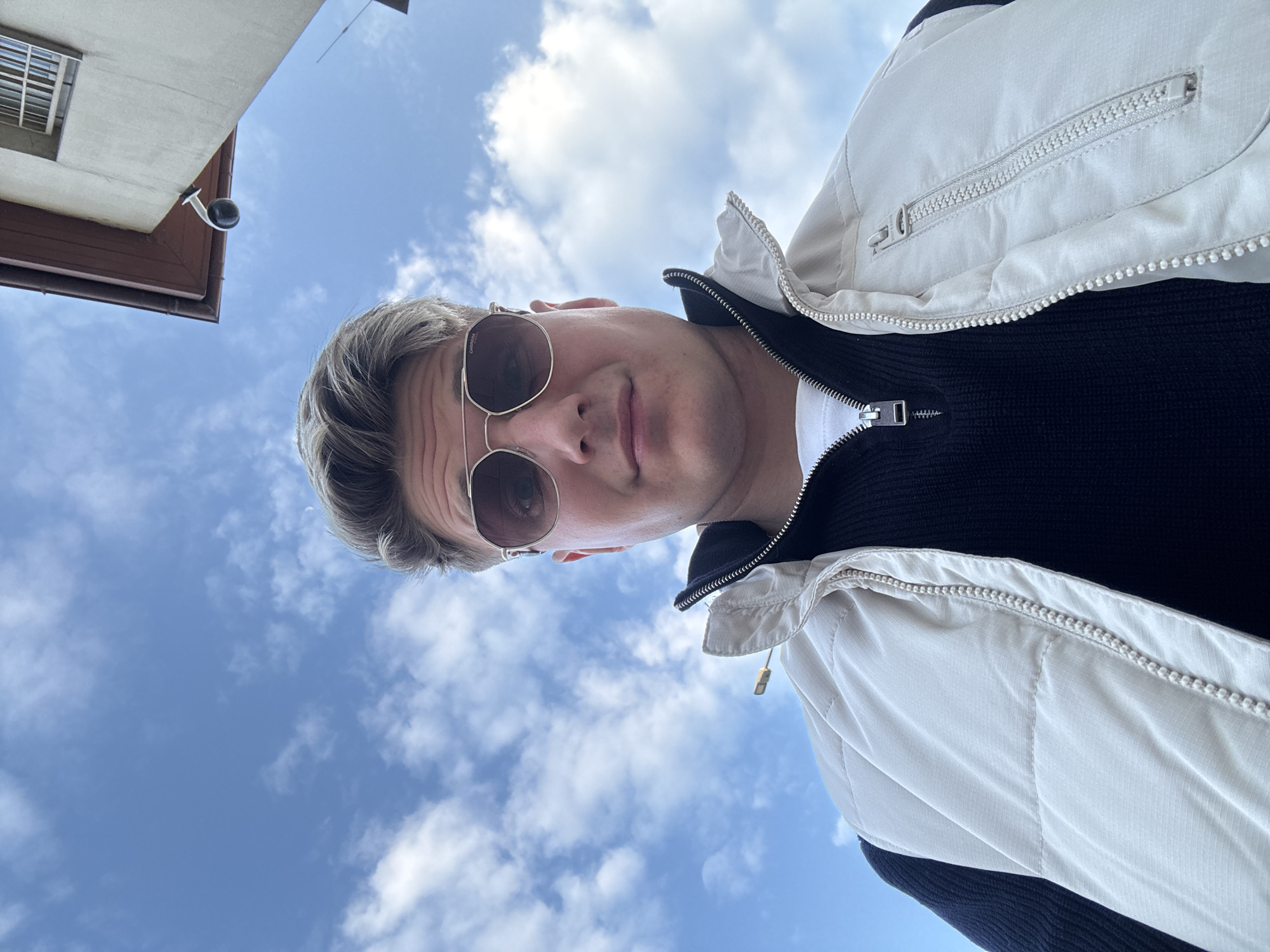 A low-angle, selfie-style photograph depicts a young adult male positioned centrally against a bright, partially cloudy blue sky and a building facade. The subject has blond hair, styled with volume, and wears aviator-style sunglasses with light brown lenses and metallic frames, bearing the "CARRERA" brand on the left lens. His expression is neutral, with a direct gaze towards the camera. He is dressed in a white crew-neck t-shirt, a black ribbed quarter-zip sweater, and a light beige puffer vest with a visible front zipper.
In the upper right background, the corner of a light-colored building is visible, featuring dark brown eaves and a partially seen window with horizontal bars. A black dome-shaped security camera is mounted on a light-colored arm beneath the building's eaves. A thin, dark rod or antenna-like object is visible against the sky on the far right. The scene is illuminated by natural daylight. This scene is located in the city of Plovdiv, Bulgaria.