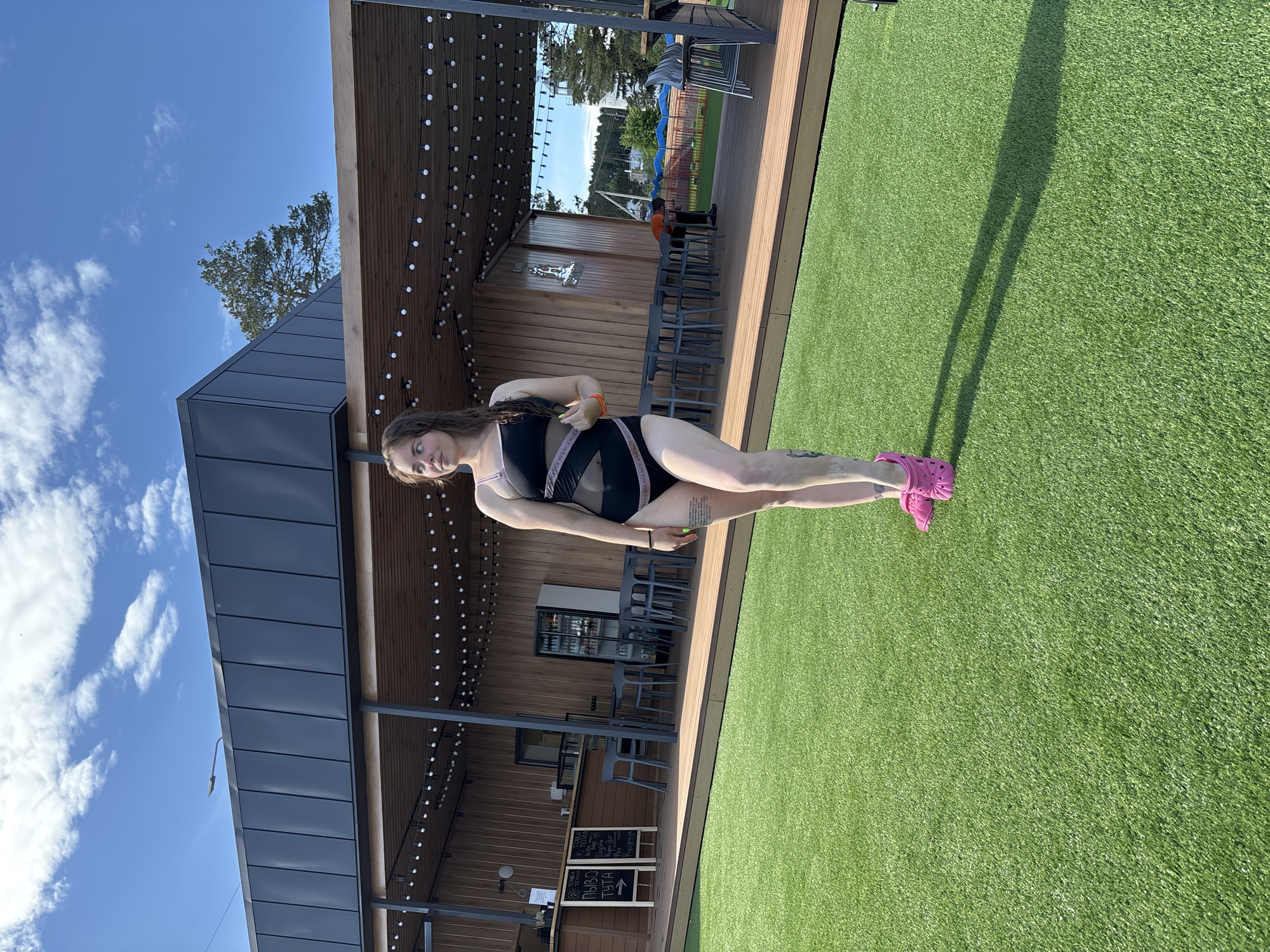 The image depicts a woman in a black one-piece swimsuit with pink trim standing on artificial turf. She is wearing pink Crocs. Behind her is a wooden building with string lights and a modern dark gray roof. There are tables and chairs under the building's covered area, and a chalkboard menu is visible near the base. The sky is blue with scattered clouds. Based on visual cues and general knowledge, the location is consistent with a recreational or tourist area, possibly near a pool or outdoor dining space. Votkinsk, Russia is the likely location, considering the prompt. No additional information about the city is discernible from the image itself.