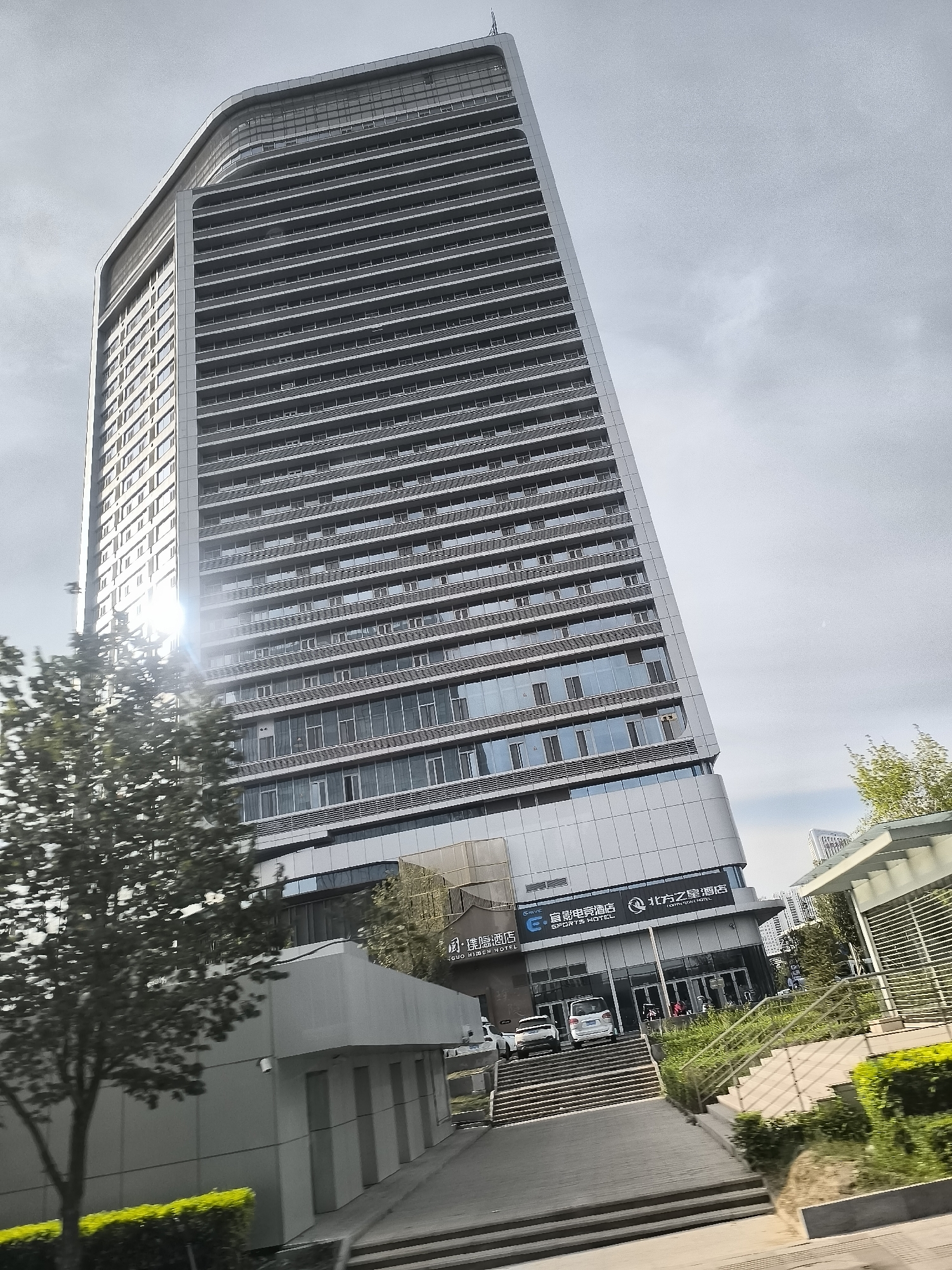 The image captures a modern, multi-story building with a distinctive curved facade, located in Ürümqi, China. The building, likely an office or hotel, is predominantly glass and metal, featuring horizontal bands of windows and metallic paneling. A bright sun glare emanates from the left side of the building, obscuring some of the detail on that section. In the foreground, a tree with dark green foliage is positioned to the left, partially obscuring the lower portion of the building. To the right of the building, a set of stairs leads up to an entrance, flanked by manicured green bushes. Several vehicles are visible near the entrance, including two white SUVs. A sign with Chinese characters and the words "SPORTS HOTEL" is present on the building facade. The sky is overcast with visible cloud formations, suggesting a diffused light condition. The overall scene depicts an urban environment with contemporary architecture.