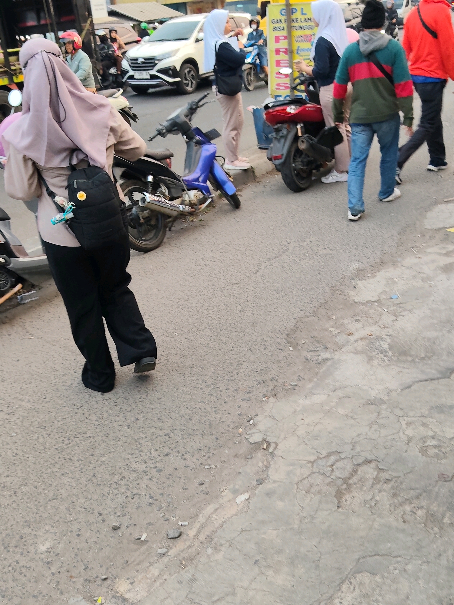 The image depicts a street scene in Purwakarta, Indonesia. In the foreground, a woman wearing a hijab and black pants is walking away from the camera, carrying a black backpack. She is standing next to a parked motorcycle. Behind her, several other people are visible, including a person in a red jacket and another in a green and red striped hoodie. A white car is parked further down the street, and other motorcycles and vehicles are present in the background. The road surface is asphalt and appears somewhat worn. The overall impression is of a busy urban environment.