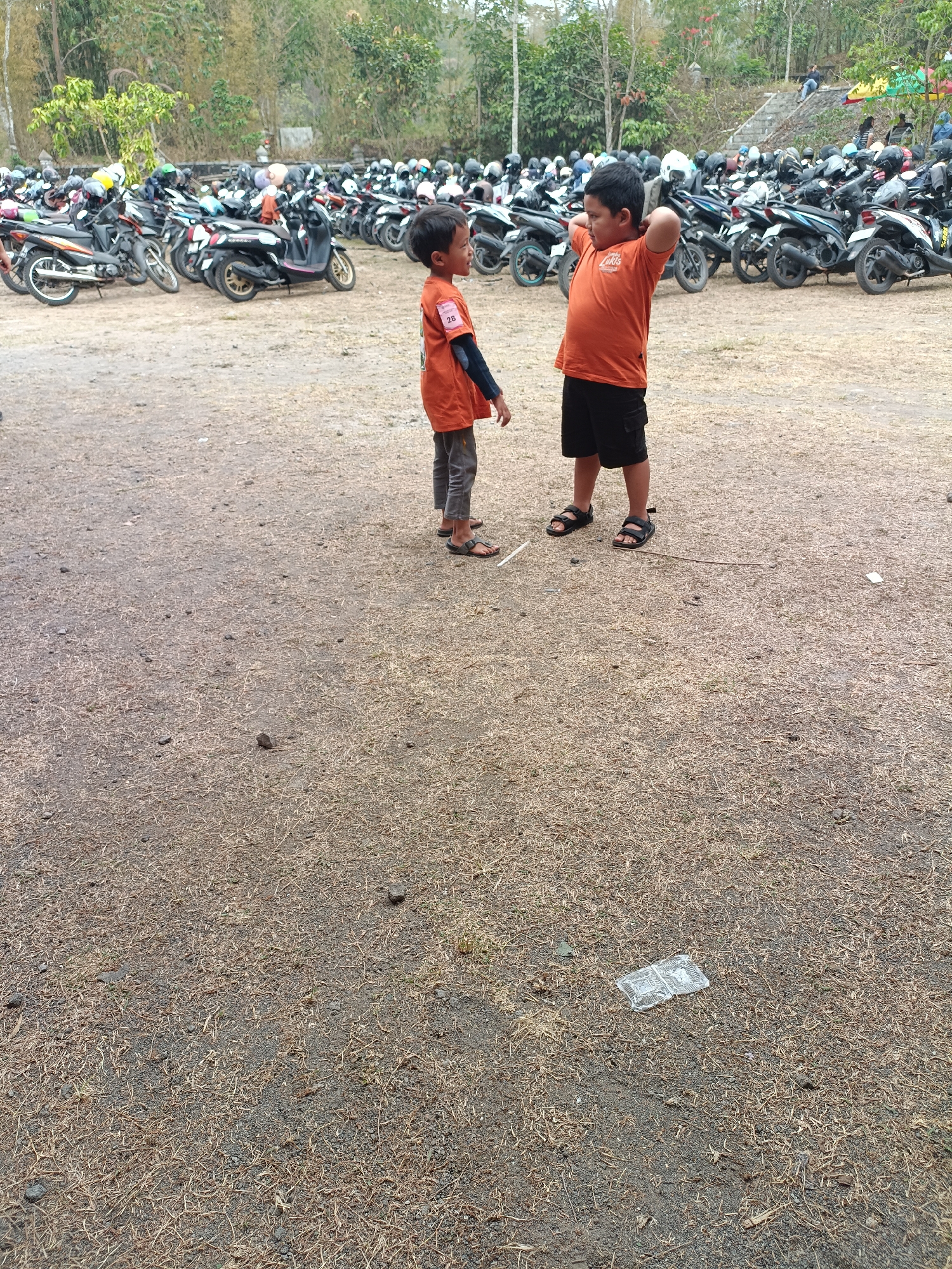 Two young boys stand in a dirt lot filled with parked motorcycles. The boy on the left wears an orange t-shirt with a white number "26" on the sleeve and dark pants. He is looking to his right. The boy on the right wears an orange t-shirt and black shorts. He is stretching his arms behind his head, looking to his left. In the background, rows of motorcycles are parked, and some trees and foliage are visible. The ground is covered in dry grass and dirt.