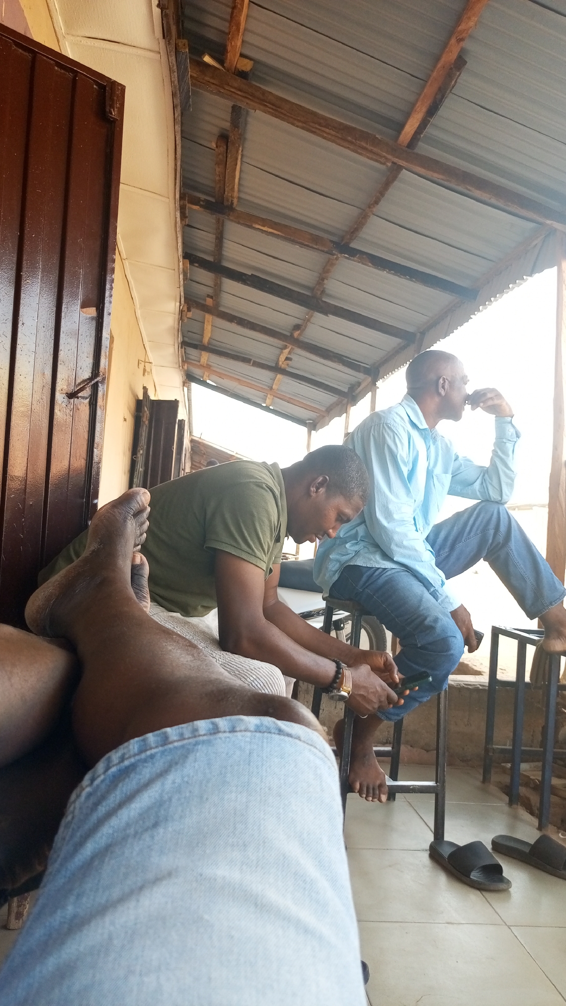 The image depicts an outdoor scene in Sabon Tasha, Nigeria. Two men are seated on metal stools under a corrugated metal roof supported by wooden beams. One man wears a light blue shirt and jeans, while the other wears a green t-shirt and jeans. The first man is leaning back on his hands, looking towards the right, while the second man is looking down at a phone in his hands. A pair of black sandals rests on the tiled floor. A brown door is visible on the left. A pair of legs and jeans partially obstruct the foreground view.