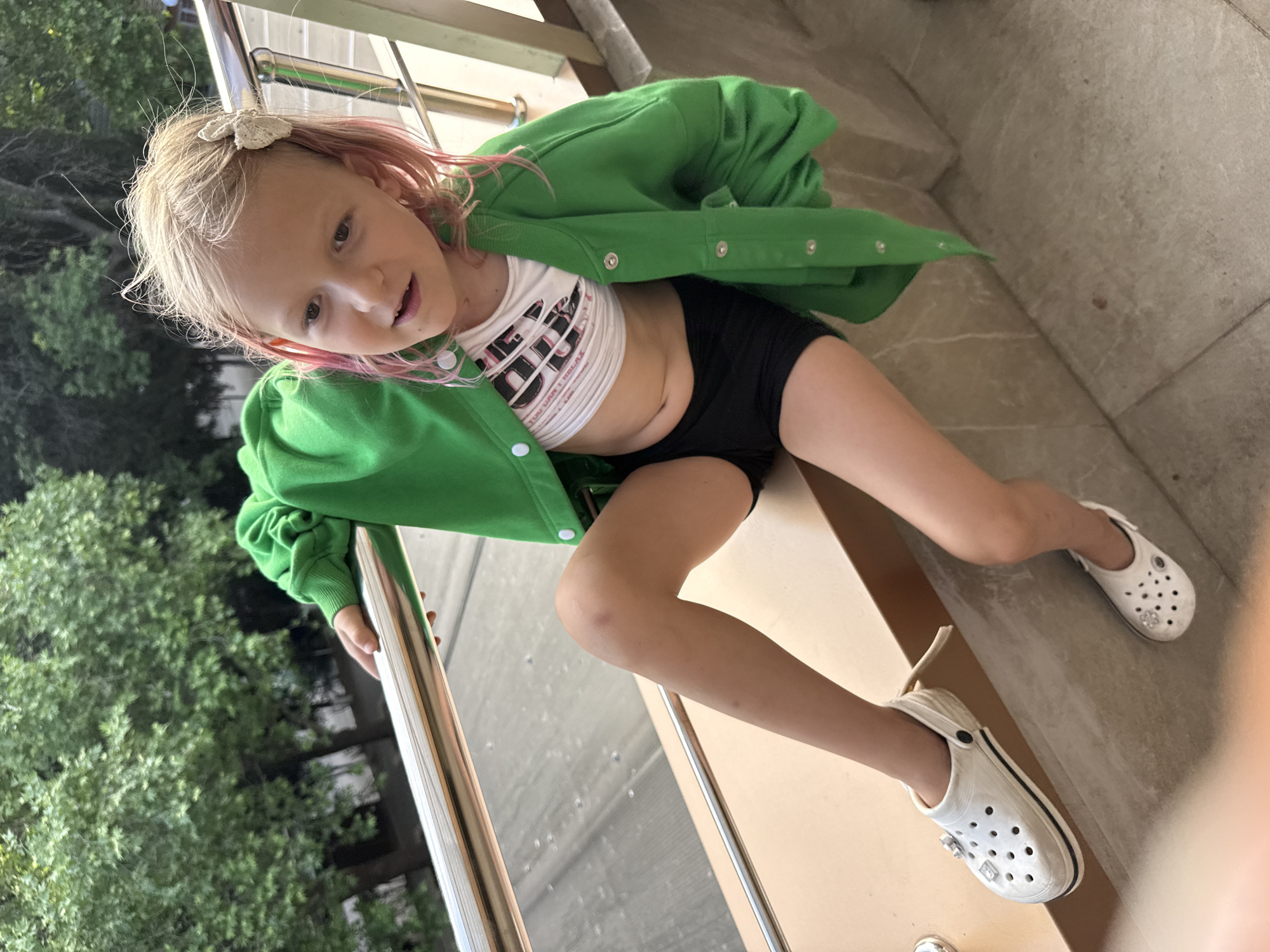 A young girl with blonde hair streaked with pink is sitting on a set of outdoor stairs. She is wearing a white crop top with a graphic print, black shorts, and white Crocs. She has a green zip-up jacket draped over her shoulders. The girl is looking up and slightly to the right, with an inquisitive expression. She has a decorative hair clip in her hair. The background consists of lush green trees and a metal railing. The lighting suggests it is daytime, possibly late afternoon. The overall mood appears casual and relaxed. No visible text is present in the image.