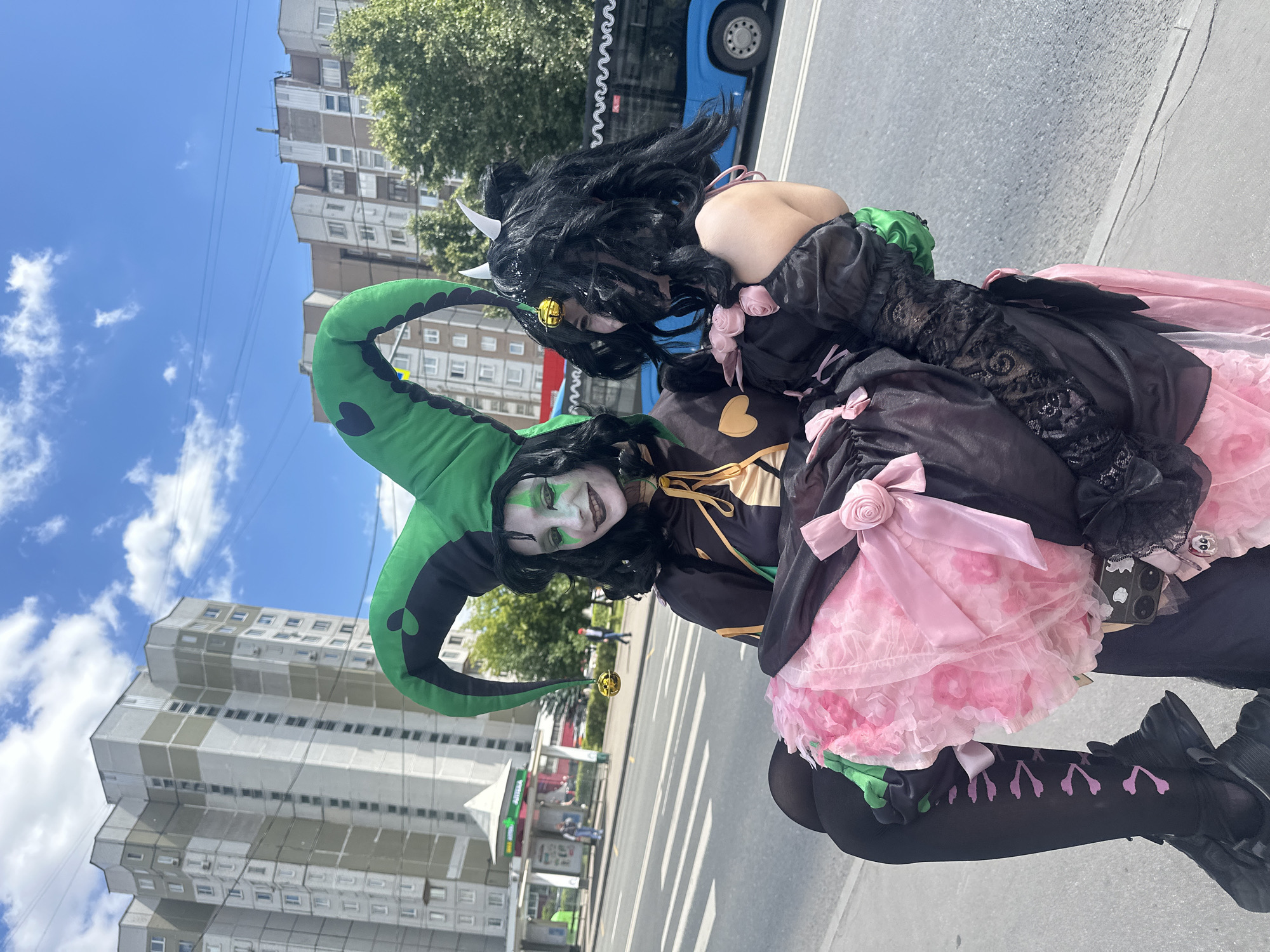 Two individuals in cosplay stand on a city street. The person on the left wears a jester's hat with green points and a black and gold outfit with face paint. They are holding the person on the right, who has black, curly hair with white horns and wears a black dress with pink ruffles. The background shows residential buildings, a bus stop, a blue bus, and a street with crosswalks. The scene appears to be in Moscow, Russia, based on the architectural style and the Cyrillic lettering on the bus stop sign.