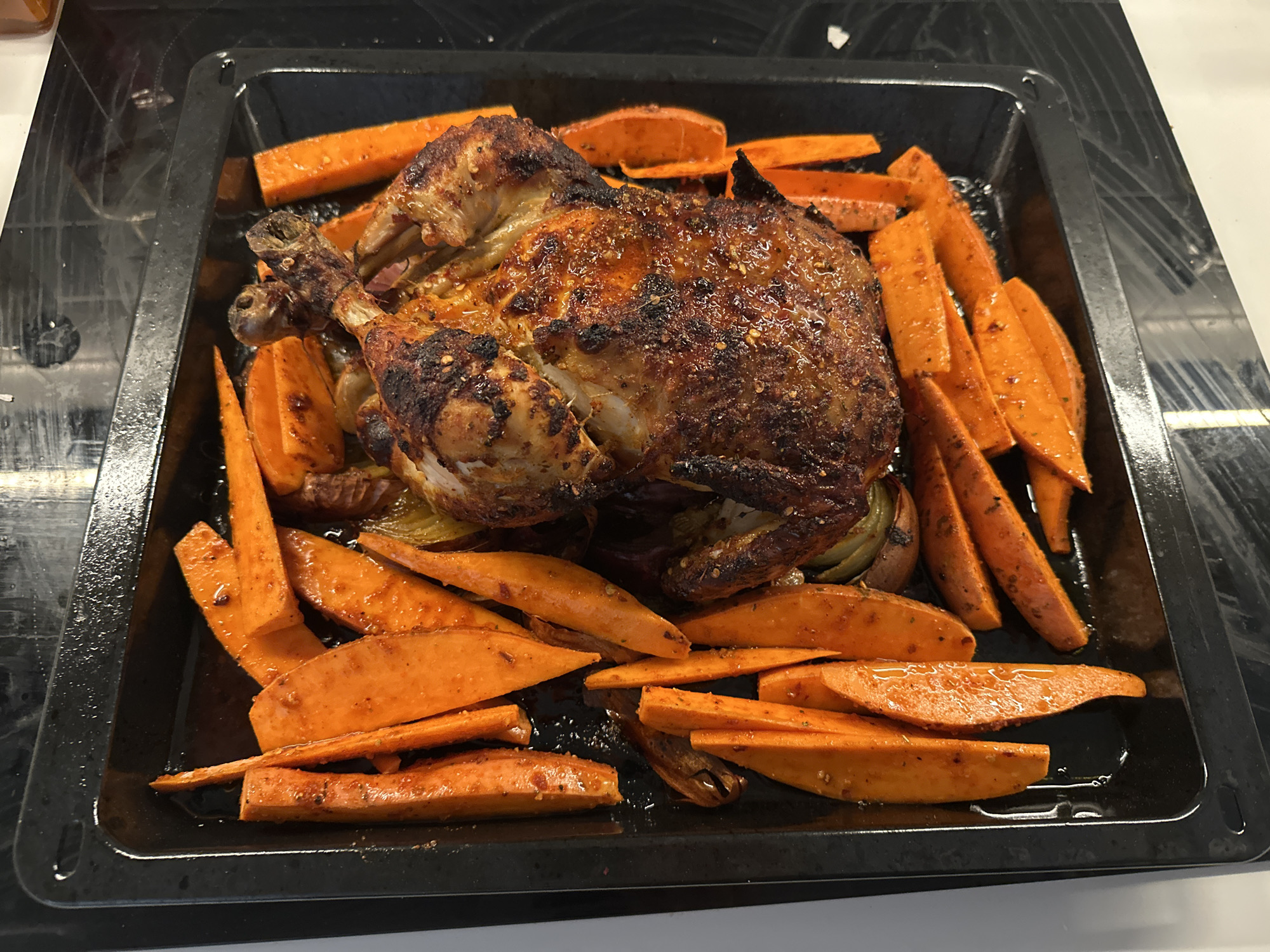 A whole roasted chicken is presented on a black baking sheet surrounded by wedges of sweet potato. The chicken has a browned, slightly charred skin and appears to be seasoned with herbs or spices. The sweet potato wedges are also browned and seasoned. Some onions are also included in the tray. The scene suggests a home-cooked meal. The precise location cannot be determined from the image, nor is there any indication that the image was taken in London.
