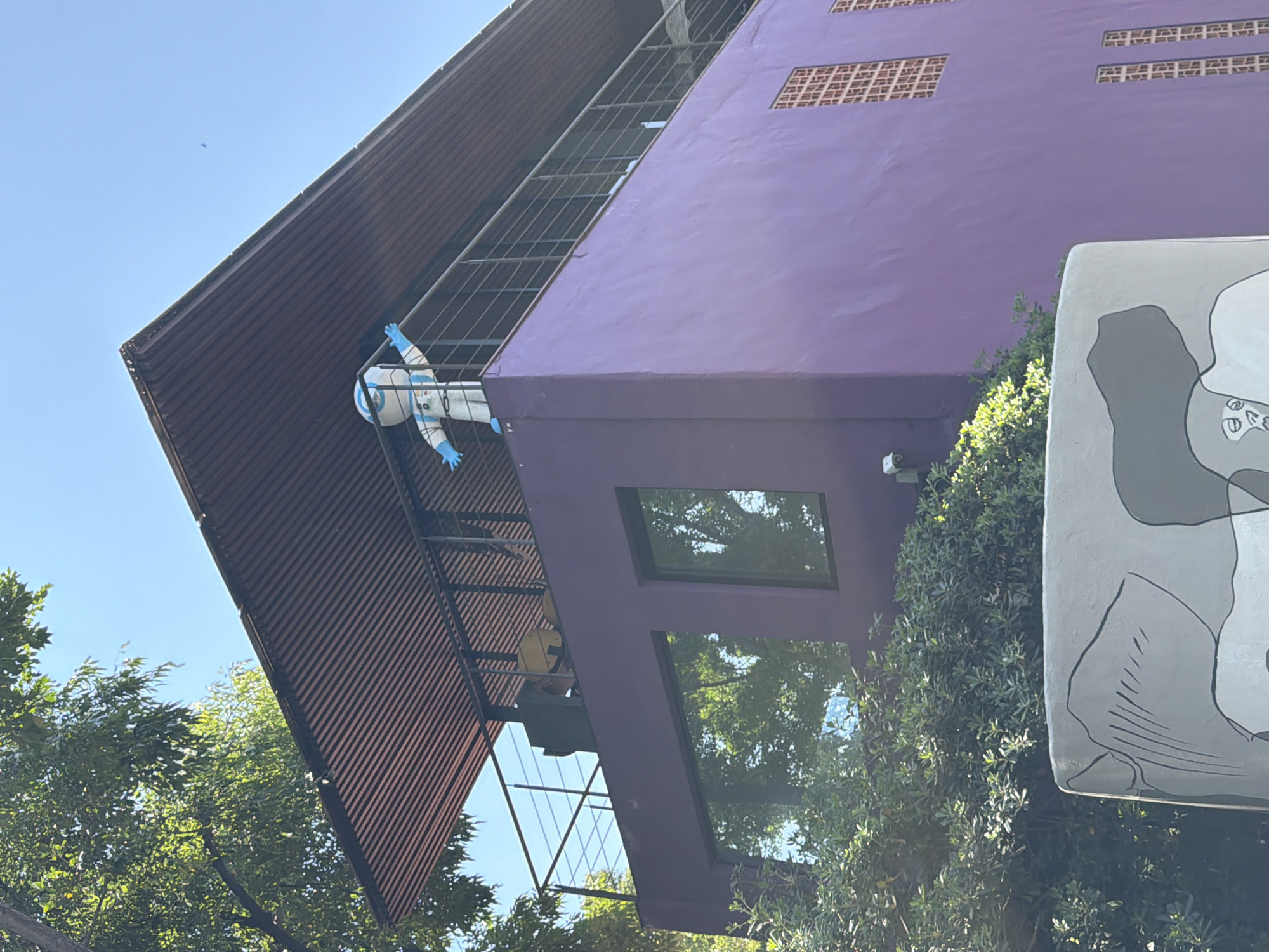The image displays a two-story building with a vibrant purple facade, located in Puebla, Mexico, under a clear blue sky. On the upper level, a large, white astronaut-like figure, featuring blue gloves and visor details, stands with outstretched arms behind a dark metal balcony railing. This upper balcony is recessed and partially covered by a wide, corrugated reddish-brown metal roof. Indistinct dark furniture is visible on the balcony behind the figure. The lower level of the building features two large rectangular windows, reflecting green foliage and the sky, alongside several smaller, lattice-patterned window openings on the right side. A white security camera is mounted on the left upper corner of the lower level. Dense green trees and shrubs surround the base of the building and frame the upper structure on the left. In the bottom foreground, a curved, light gray concrete structure partially obstructs the view, decorated with abstract black and dark gray line art. The scene is brightly illuminated by direct sunlight.