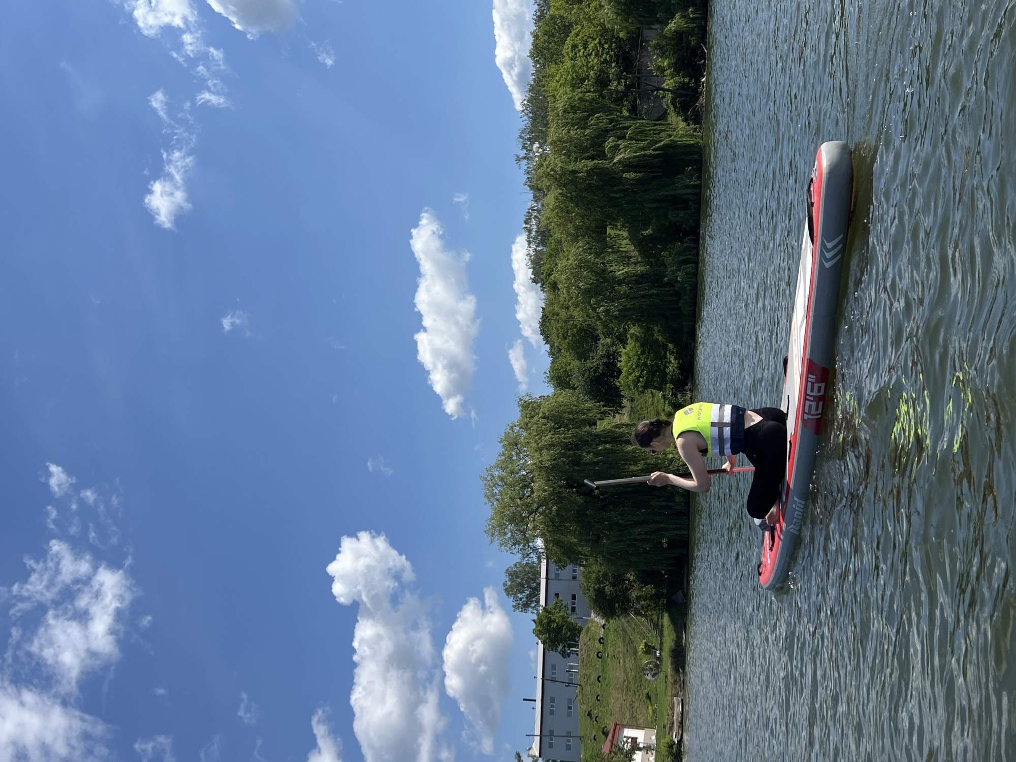 A woman is paddleboarding on a lake in Bila, Ukraine. She is wearing a yellow life vest and black leggings. She is sitting on the paddleboard, holding a paddle. The paddleboard is red and gray and has "Rapid 12'6\"" printed on it. The lake is calm, with ripples on the surface. In the background, there are green trees and a building. The sky is blue with white clouds.