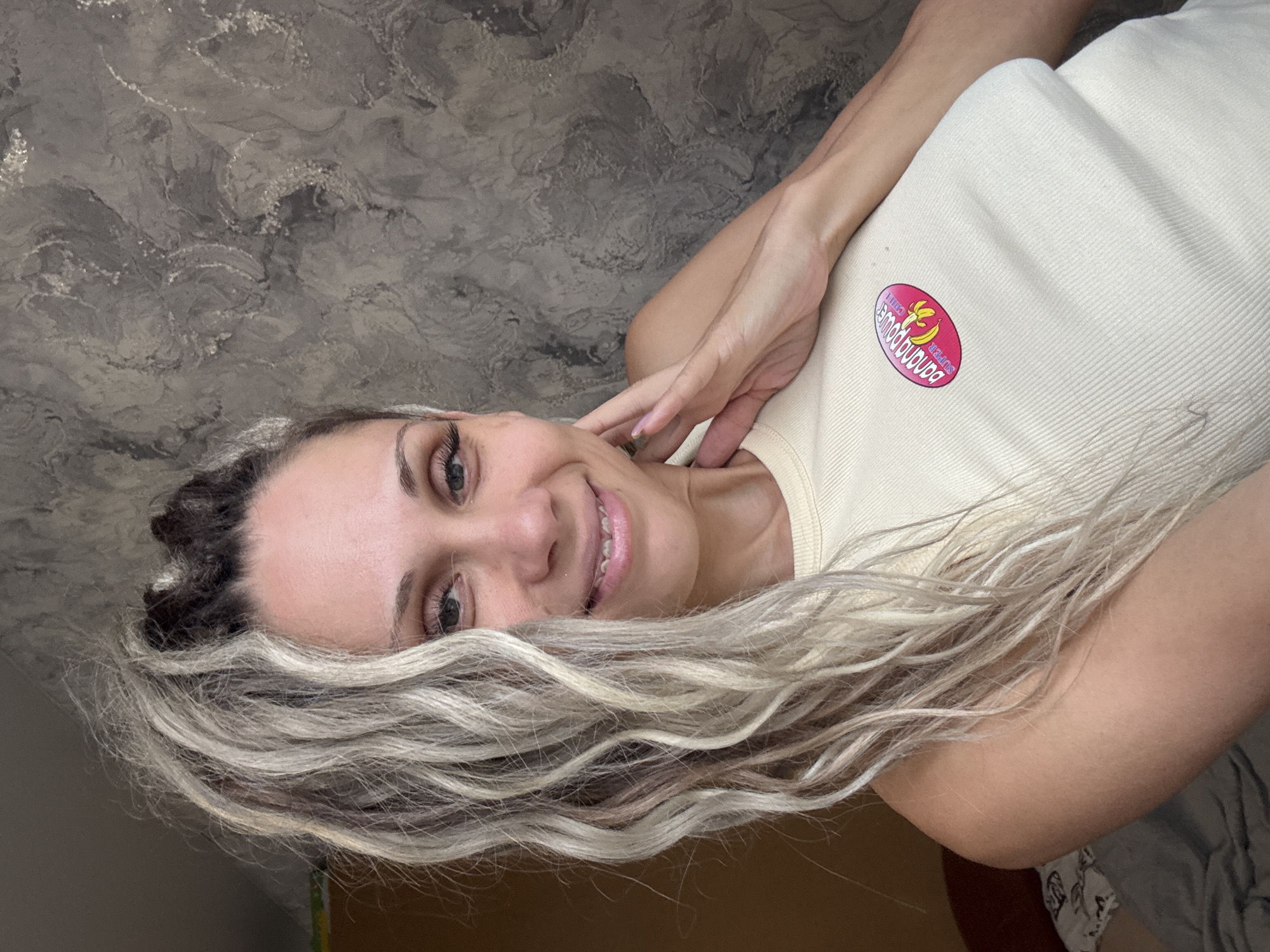 A woman with blonde and dark braided dreadlocks smiles at the camera, resting her left hand on her neck. She is wearing a light yellow ribbed tank top with a pink oval sticker featuring a banana and the words "SUPER BANANA POWER CHILL". The background is a textured grey wall. The image appears to be taken indoors, with a hint of brown wall and a bed visible on the left. There is no indication of the location being Rusanovo, Russia.