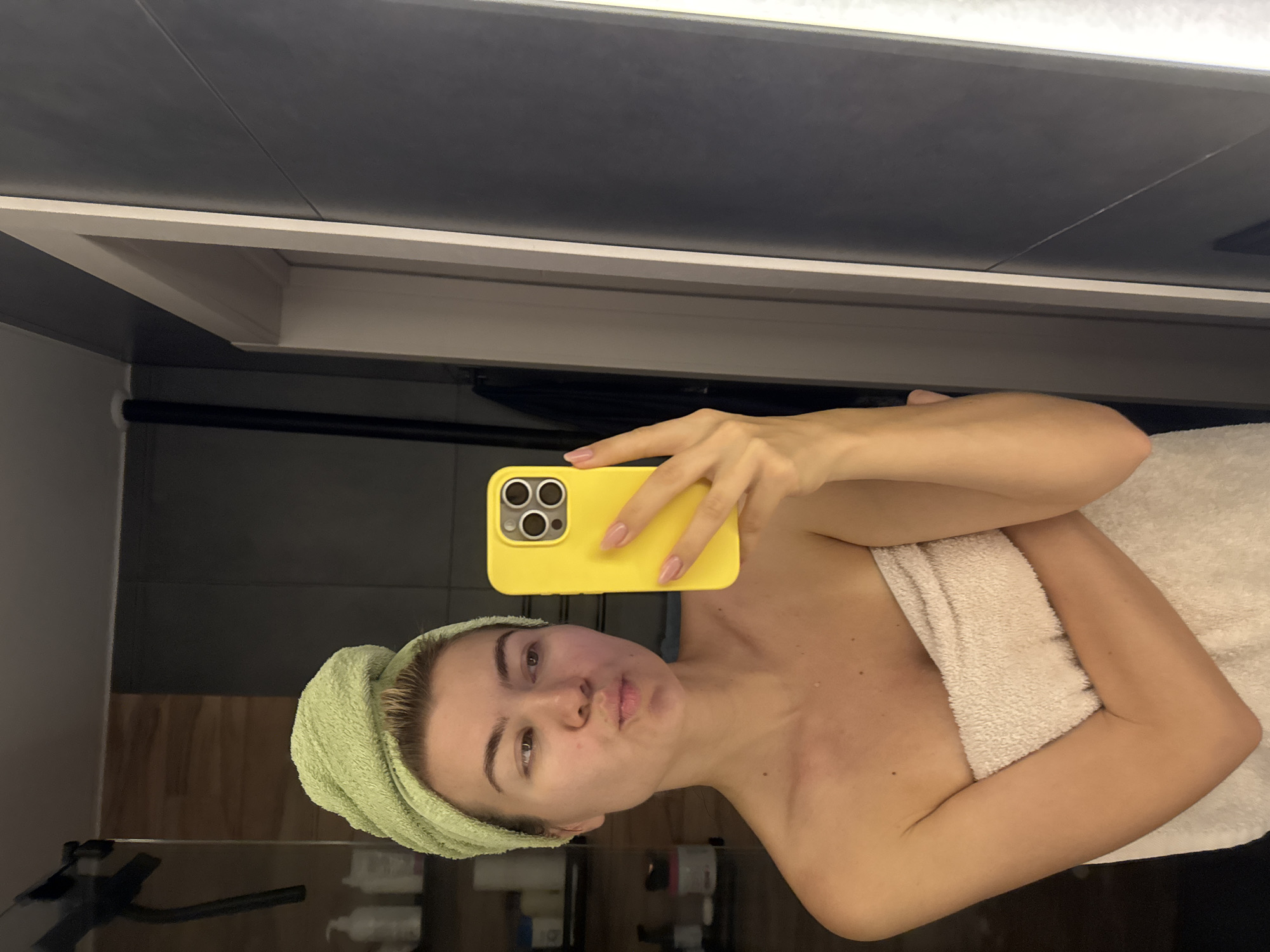 A woman is holding a yellow iPhone up to take a selfie in what appears to be a bathroom. She has a light green towel wrapped around her head and is wrapped in a white towel. In the background are bathroom walls and possibly a shelf with bottles on it. The location is Unknown, Unknown.